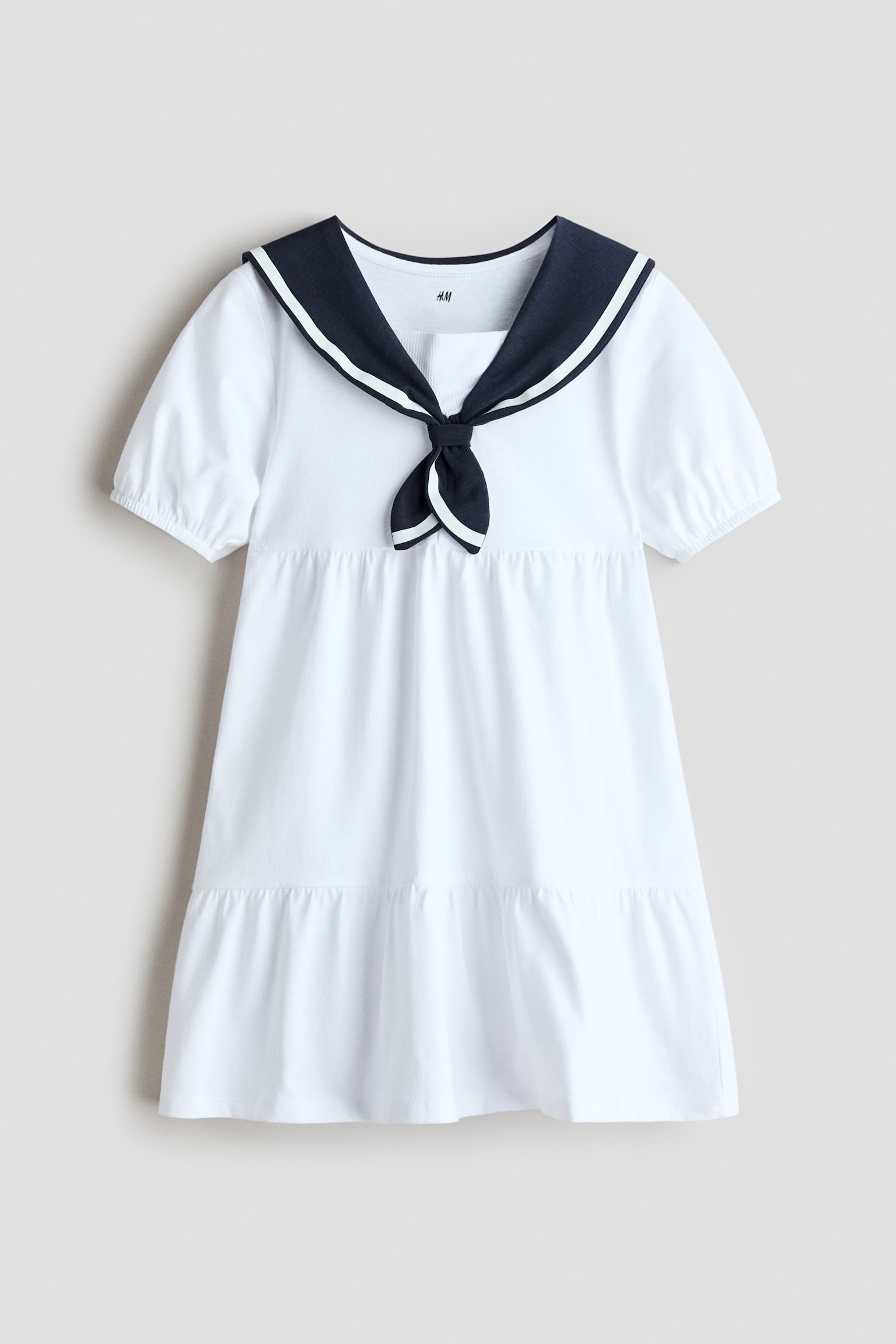 Presenting a white tiered A-line dress crafted from soft cotton jersey, showcasing its front. It features a navy sailor collar adorned with white stripes, highlighted by a decorative bow at the front, and short puff sleeves with elasticated cuffs. - 1