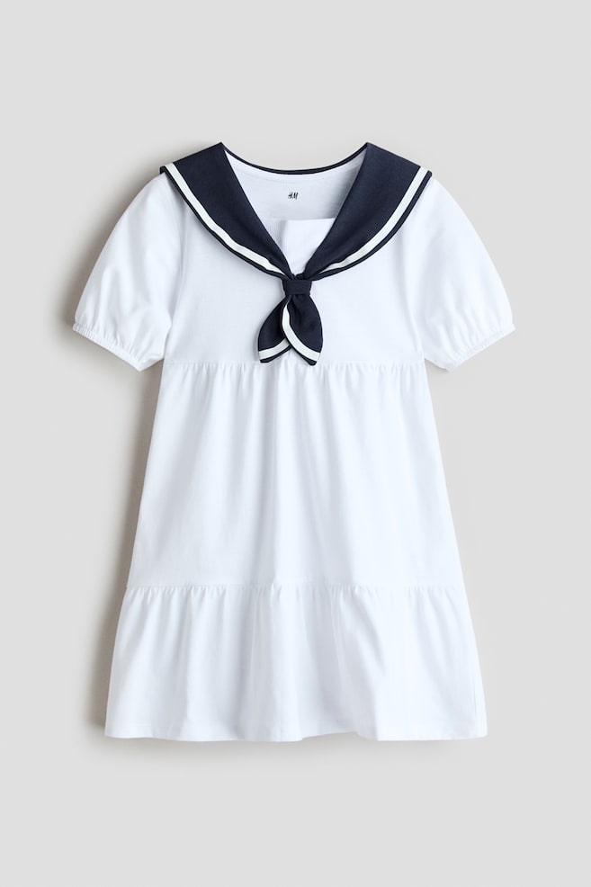 Presenting a white tiered A-line dress crafted from soft cotton jersey, showcasing its front. It features a navy sailor collar adorned with white stripes, highlighted by a decorative bow at the front, and short puff sleeves with elasticated cuffs. - 1