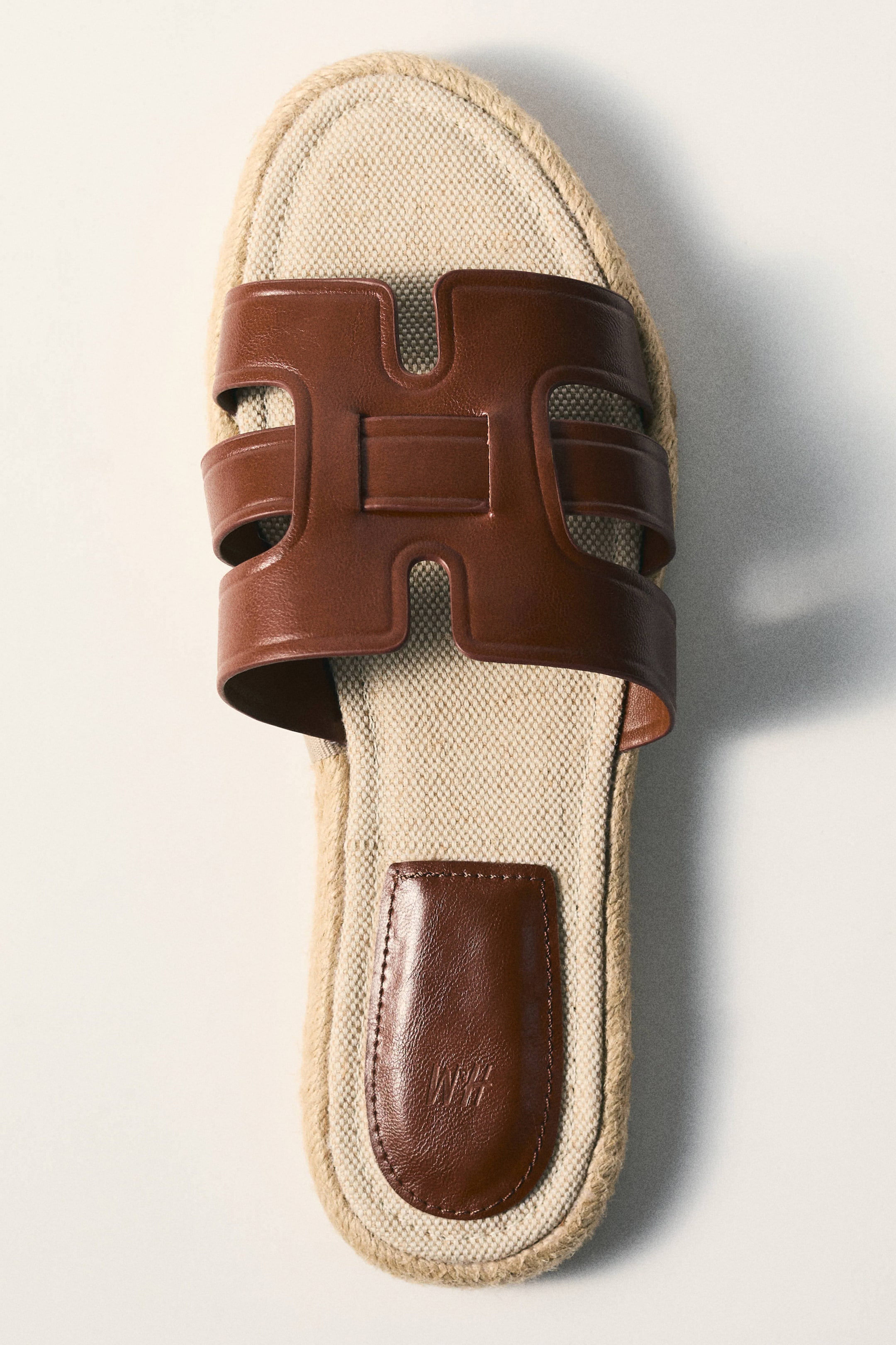 Vis større bilde: En slip-in sandal vises fra et topp-ned, lett vinklet perspektiv, som fremhever en brun lærrem som danner en H-form over vristen. Fotsengen er laget av et lys beige vevd tekstil, og sålens omkrets er innpakket med naturlig jutesnor, med en liten, avrundet brun lærdetalj på hælområdet.