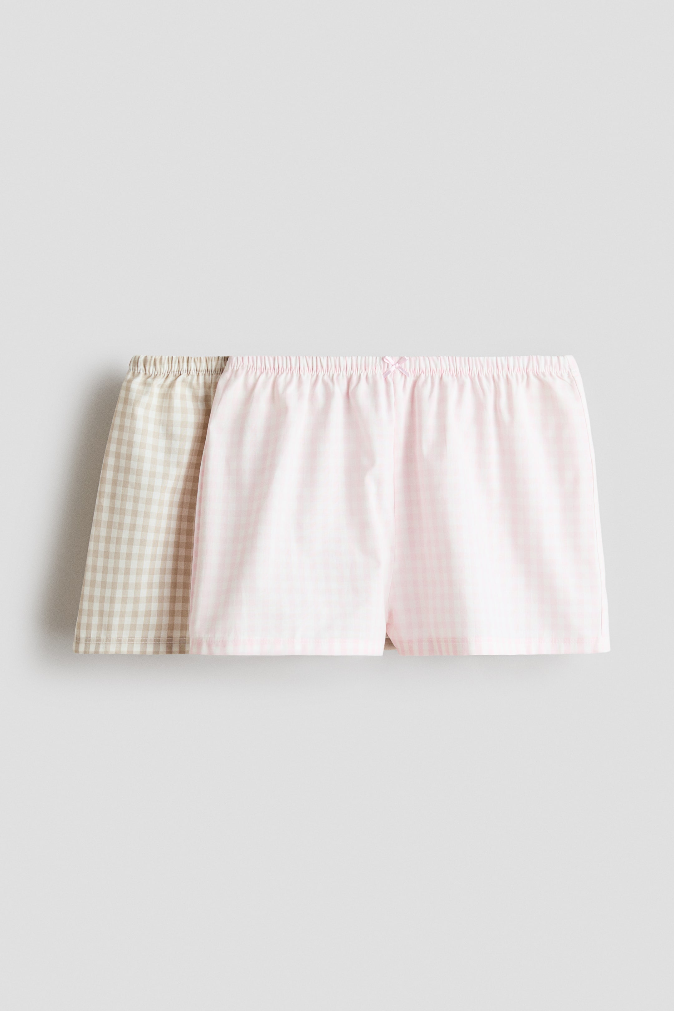 Vis større bilde: Pyjamashorts i en myk bomullsvev med elastisk snøring i midjen presenteres som et sett på to. Det fremre paret viser et lys rosa og hvitt gingham-mønster, med en liten rosa sløyfe i midten av den elastiske midjen. Et annet par, i et lys beige og hvitt gingham-mønster, er delvis synlig bak det første.