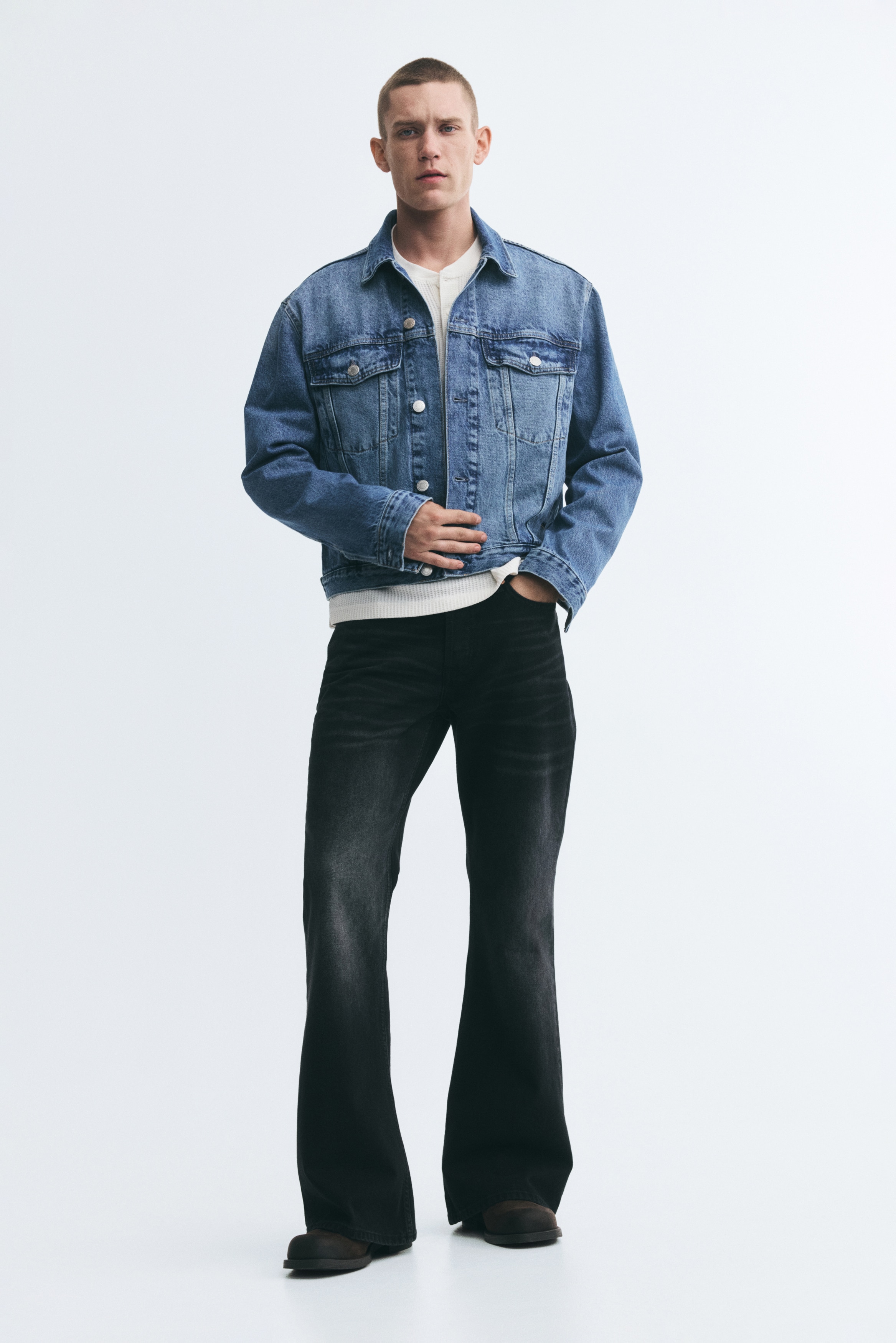 A light blue denim jacket is shown front on, featuring a classic point collar, a button-up front fastening with silver-toned metal buttons, and two buttoned chest flap pockets. The jacket also features two vertical welt side pockets, long sleeves with buttoned cuffs, and a lightly washed finish.