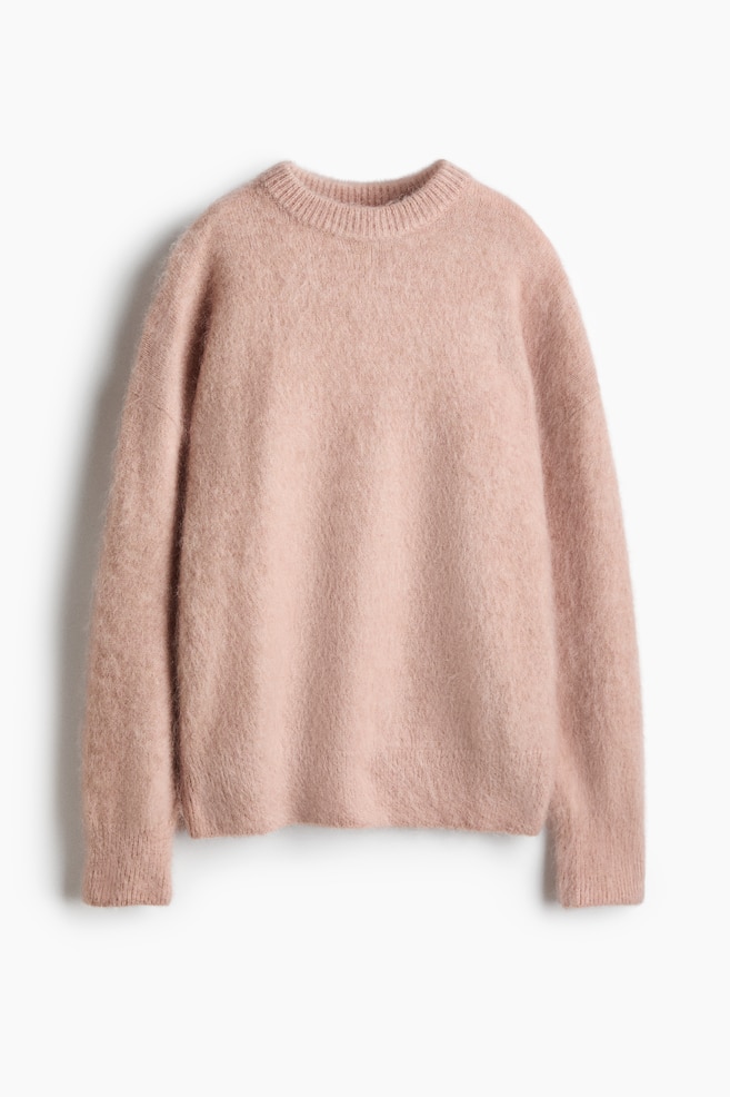 En oversized, lys rosa strikkegenser henger i frontvendt visning og viser frem den myke, lodne strukturen. Genseren har rund hals med ribbestrikket kant, senkede skuldre og lange ermer med ribbestrikkede mansjetter og ribbestrikket nederkant. - 1