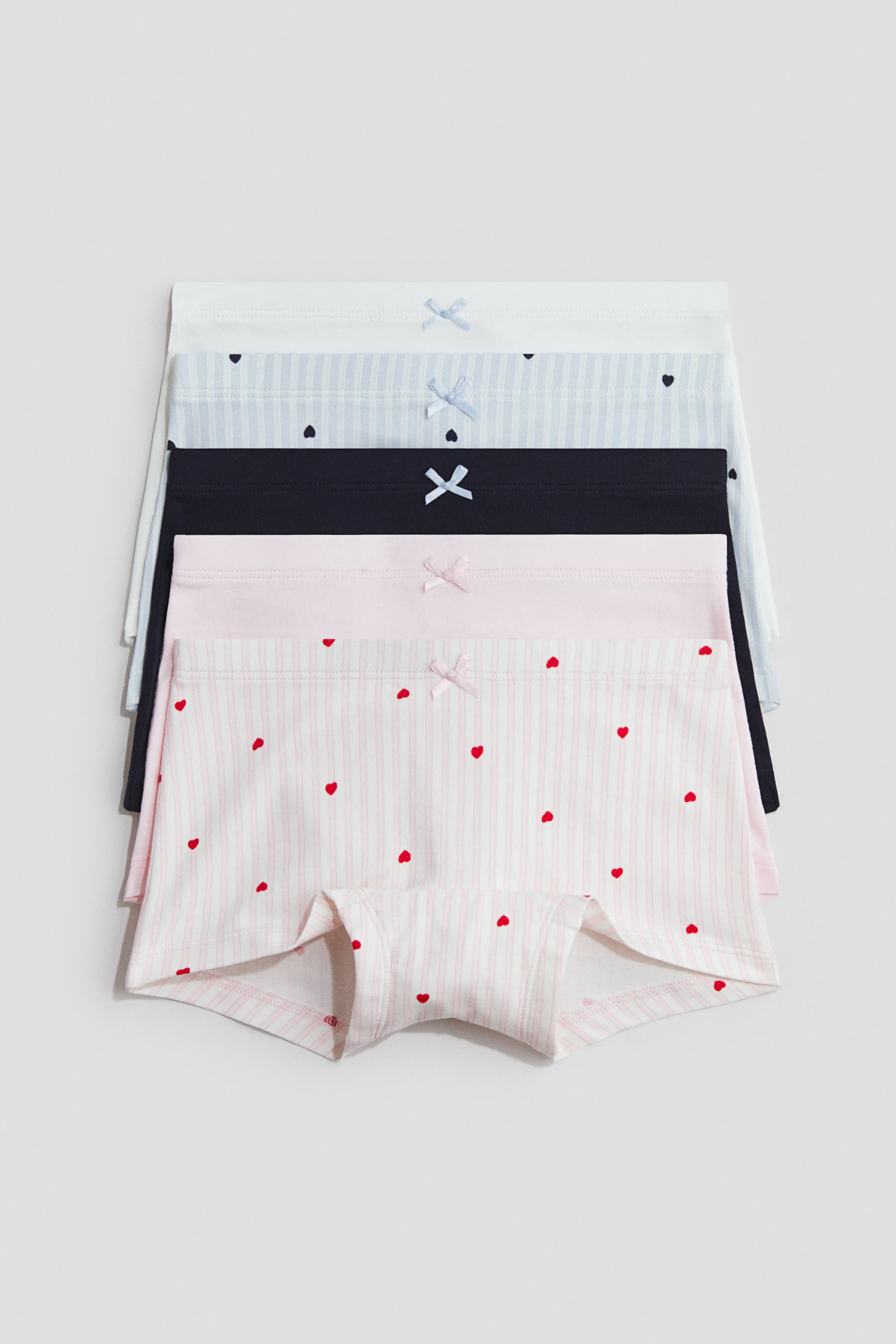 Vis større bilde: Fem par boksershorts i myk bomullsjersey er stablet, hver med en elastisk midje og en liten sløyfe foran. Stabelen inkluderer et hvitt par, et lyseblått stripete par med mørkeblå hjerter, et ensfarget mørkeblått par, et ensfarget lyserosa par, og et lyserosa stripete par med røde hjerter, hvor det nederste paret viser benåpningen fra en frontvendt vinkel.