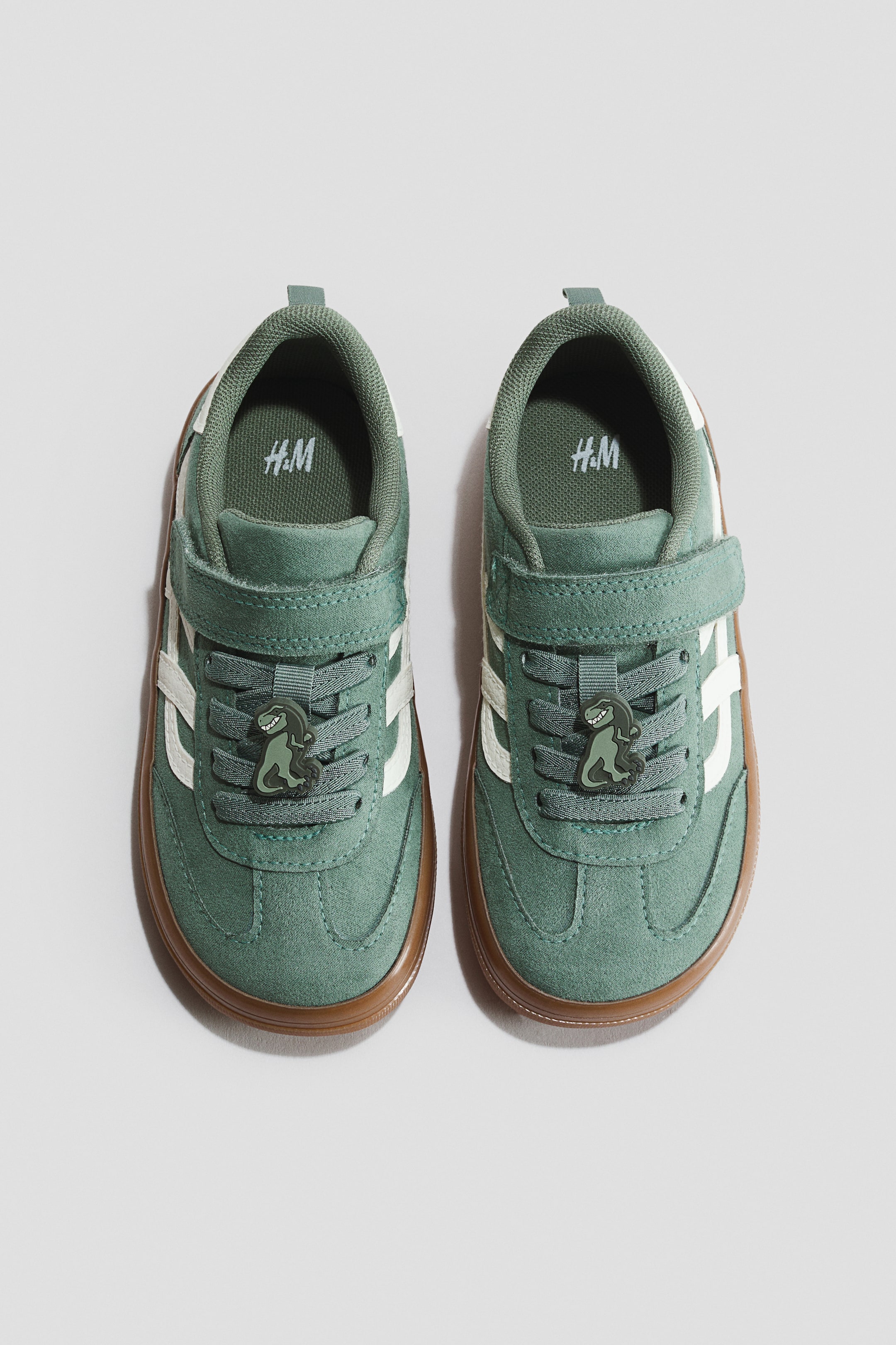 View larger image: A pair of green sneakers is shown from the front, featuring elastic laces with a dinosaur charm and a hook-and-loop tab closure. White stripes detail the sides, and a brown rubber sole is visible.