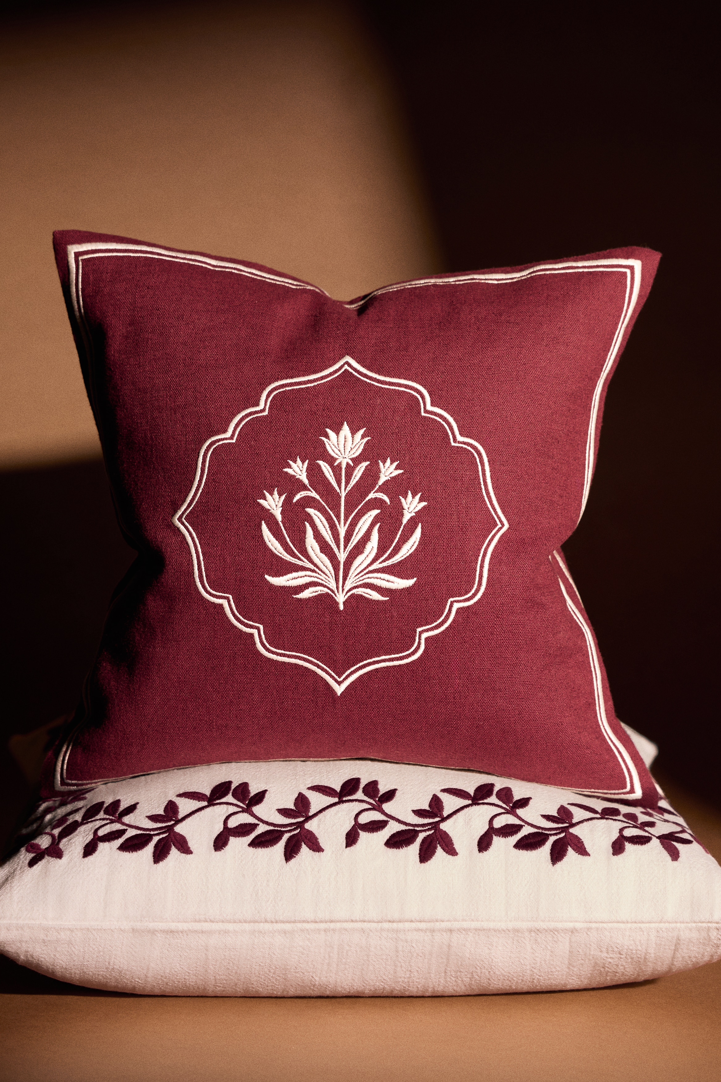 A square burgundy cushion is shown front-on, featuring a central white embroidered floral motif enclosed within a scalloped frame. A thin, double-lined white embroidered border frames the outer edges of the cushion.