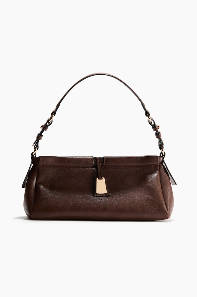 A dark brown shoulder bag is showcased in its front view, highlighting a sleek texture and a slim, adjustable strap adorned with gold-toned hardware. A rectangular gold-toned charm elegantly dangles from the central flap closure. - 1