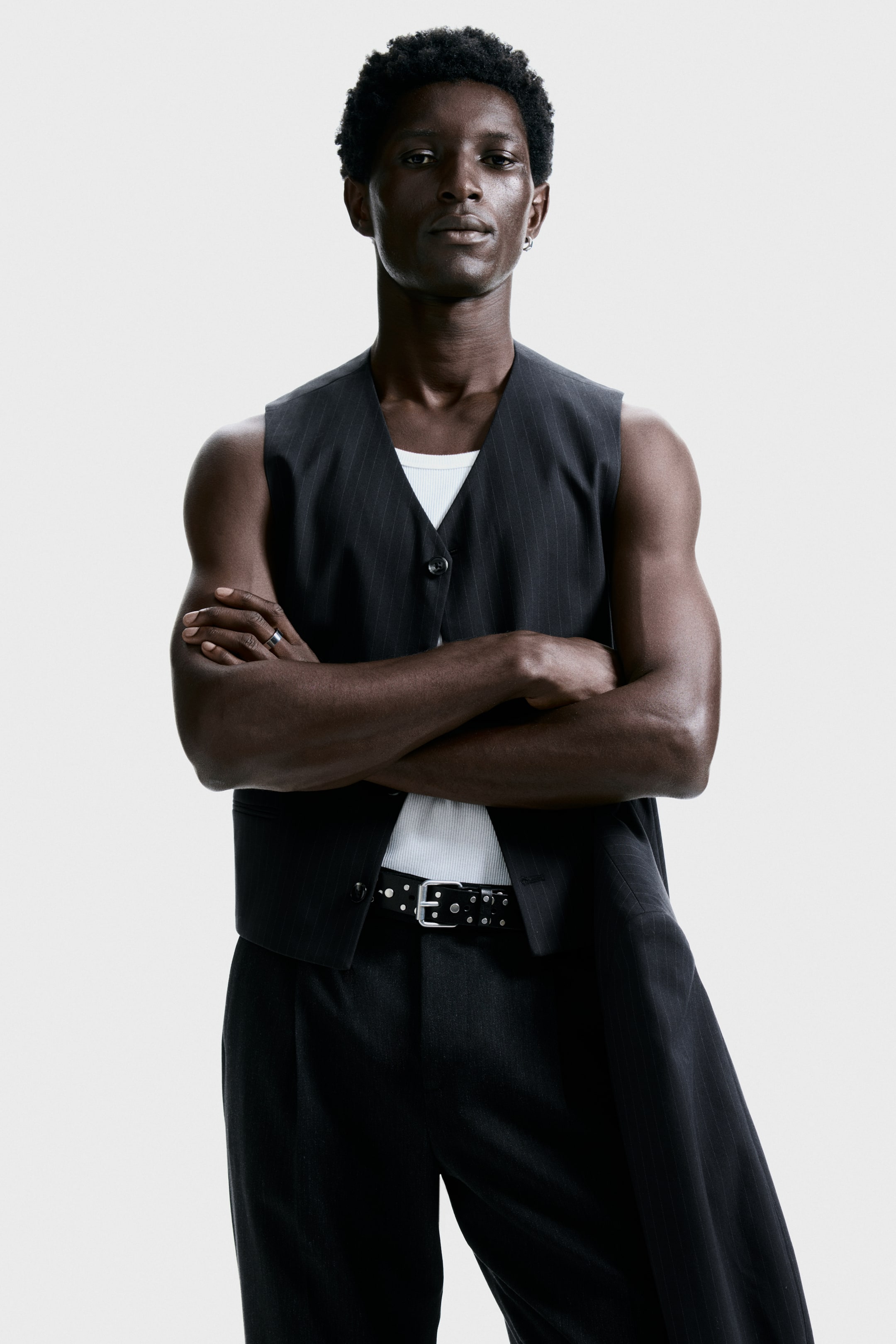 View larger image: A dark pinstriped vest and matching pleated trousers are being presented from a frontal angle. The vest is featuring a V-neck and button-front closure, layering over a white ribbed tank top, and the trousers are styled with a black studded belt.