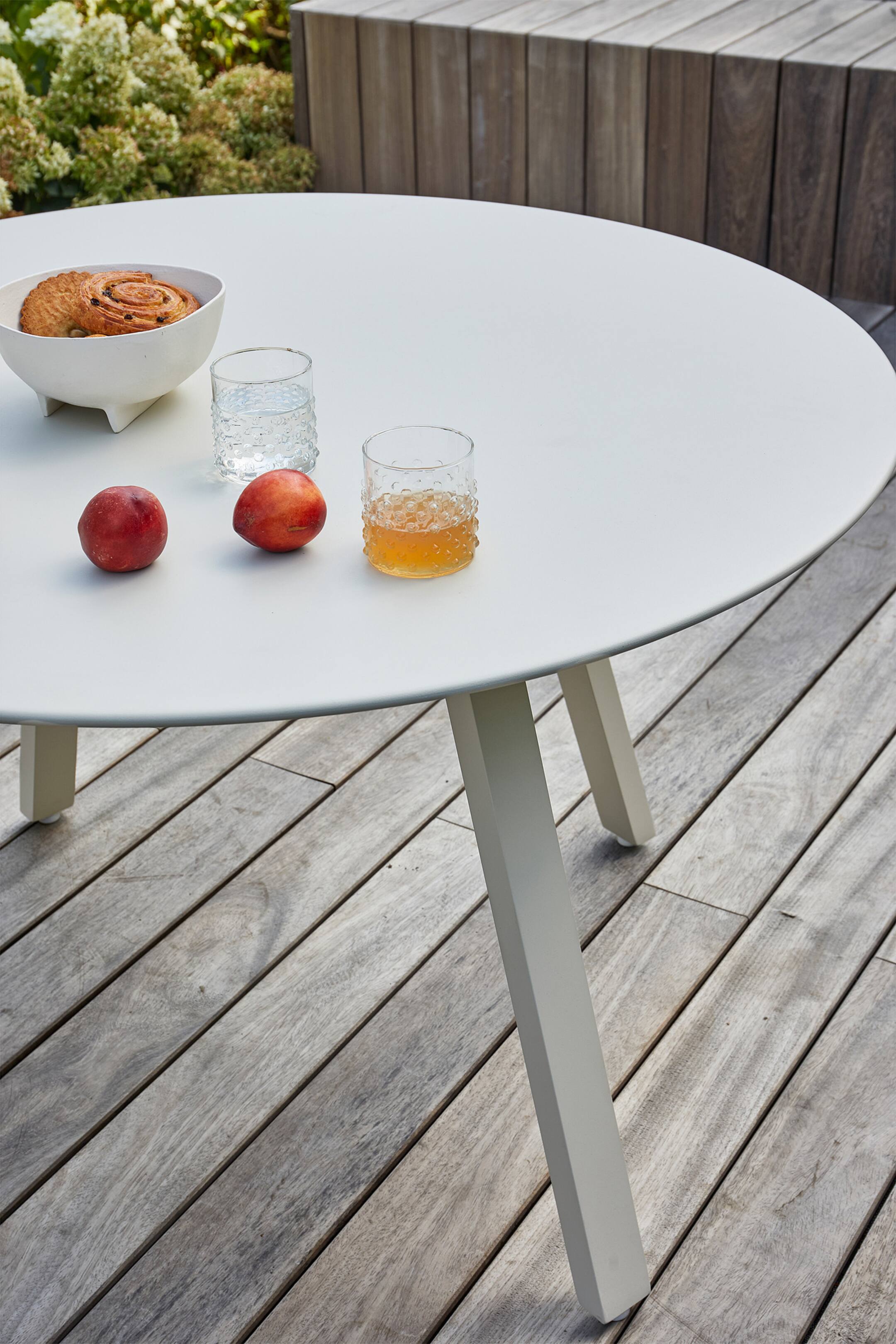 Agrandir l'image: Une table d'extérieur ronde de couleur gris clair est présentée en hauteur, avec un plateau lisse et mat, et trois pieds rectangulaires visibles qui sont inclinés vers l'extérieur. Divers objets, dont un bol blanc avec des pâtisseries, deux verres transparents et deux pêches, reposent sur sa surface.