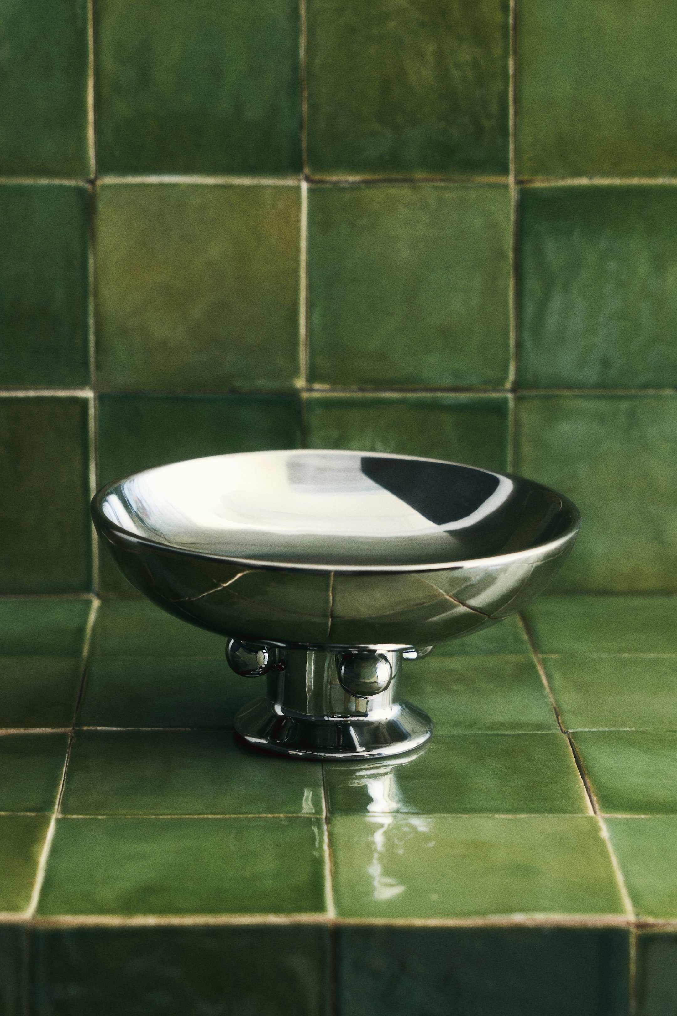 View larger image: A footed bowl in electroplated stoneware with a metallic finish is presenting its front. It features a wide, shallow basin and a flared base, with three spherical details adorning the stem.