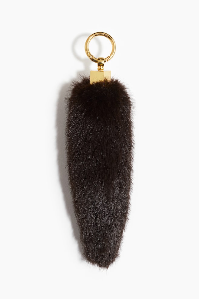 A dark brown fur key fob is featuring an elongated, tapering body. A gold-toned metal ring is resting atop a rectangular connector, which is securing the fur fob at its upper end. - 1