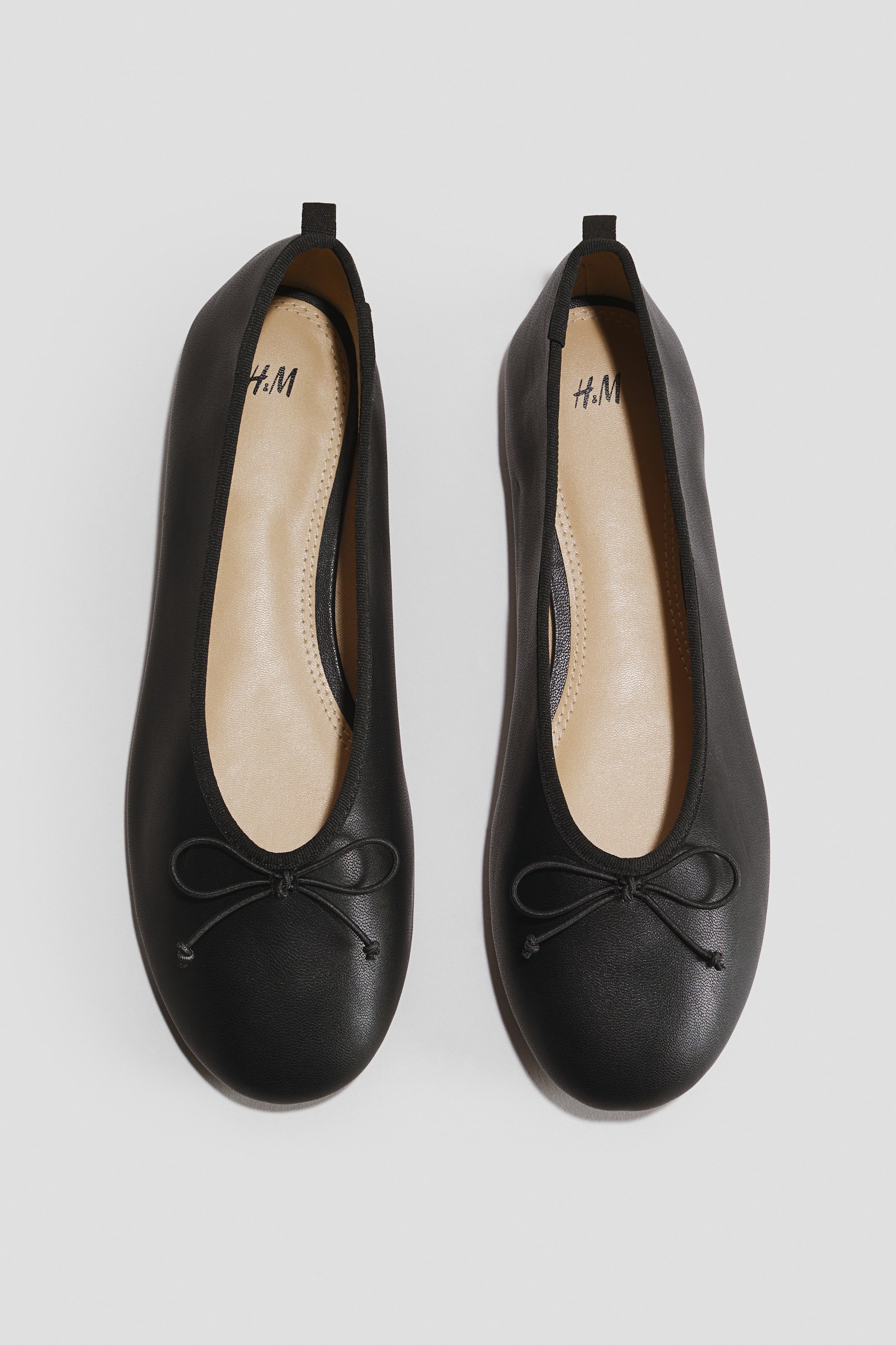 Visualizza immagine più grande: Due ballerine nere presentano una punta arrotondata, un piccolo fiocco decorativo sulla tomaia anteriore e un bordo superiore elasticizzato. Ogni scarpa mostra una soletta interna beige chiaro con il logo "H.M" e un tirante nero sul tallone posteriore.