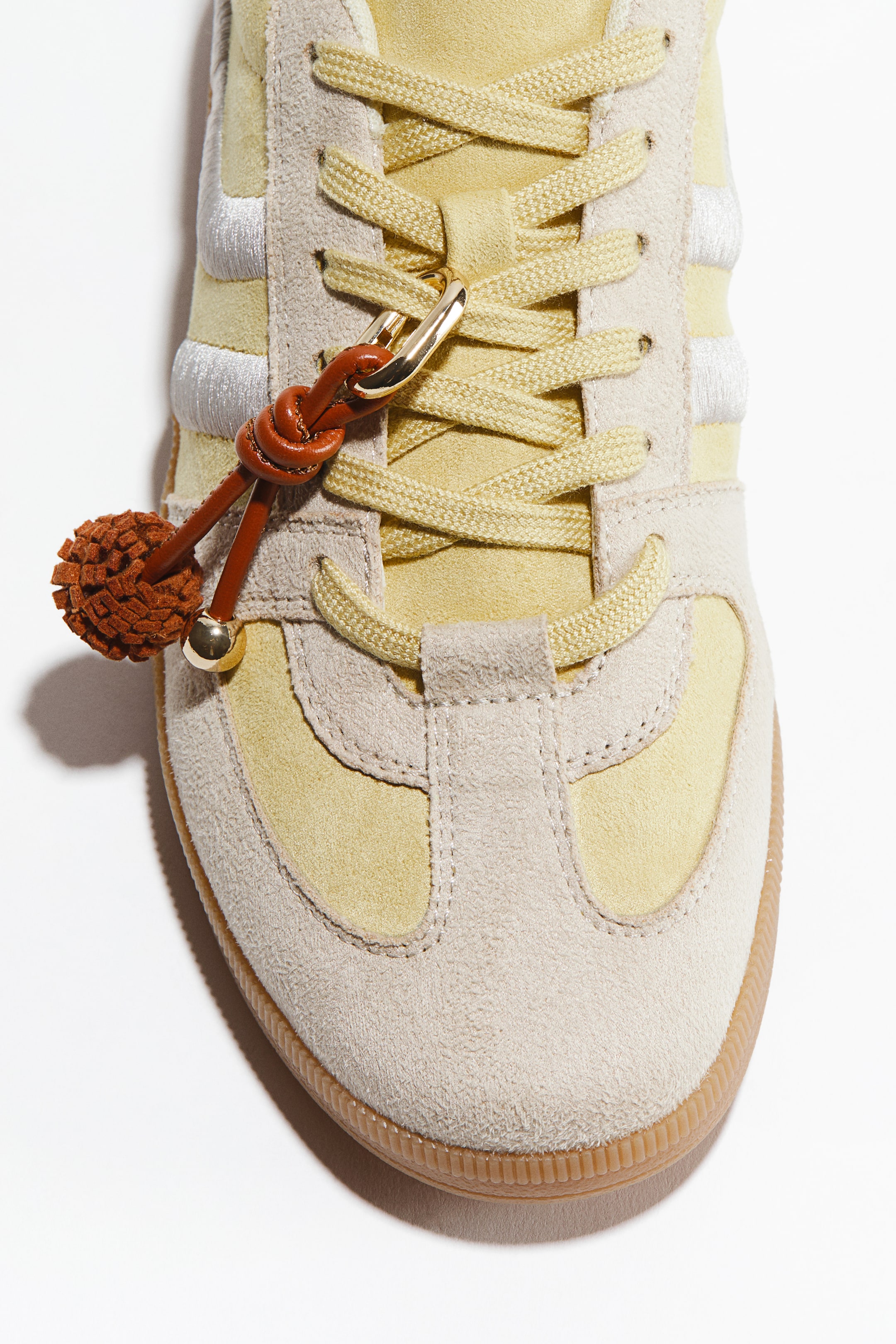 Zobacz większy obraz: But w pl-PL The sneaker is featuring a light yellow suede upper with light beige suede overlays, facing front, and flat yellow laces are crisscrossing through its eyelets. Brązowy skórzany charm z plecionym wzorem, złotym klipsem i teksturowaną kulistą pomponem jest przymocowany do sznurowadeł, podczas gdy trzy białe haftowane paski zdobią bok, a żebrowana gumowa podeszwa otacza dół.