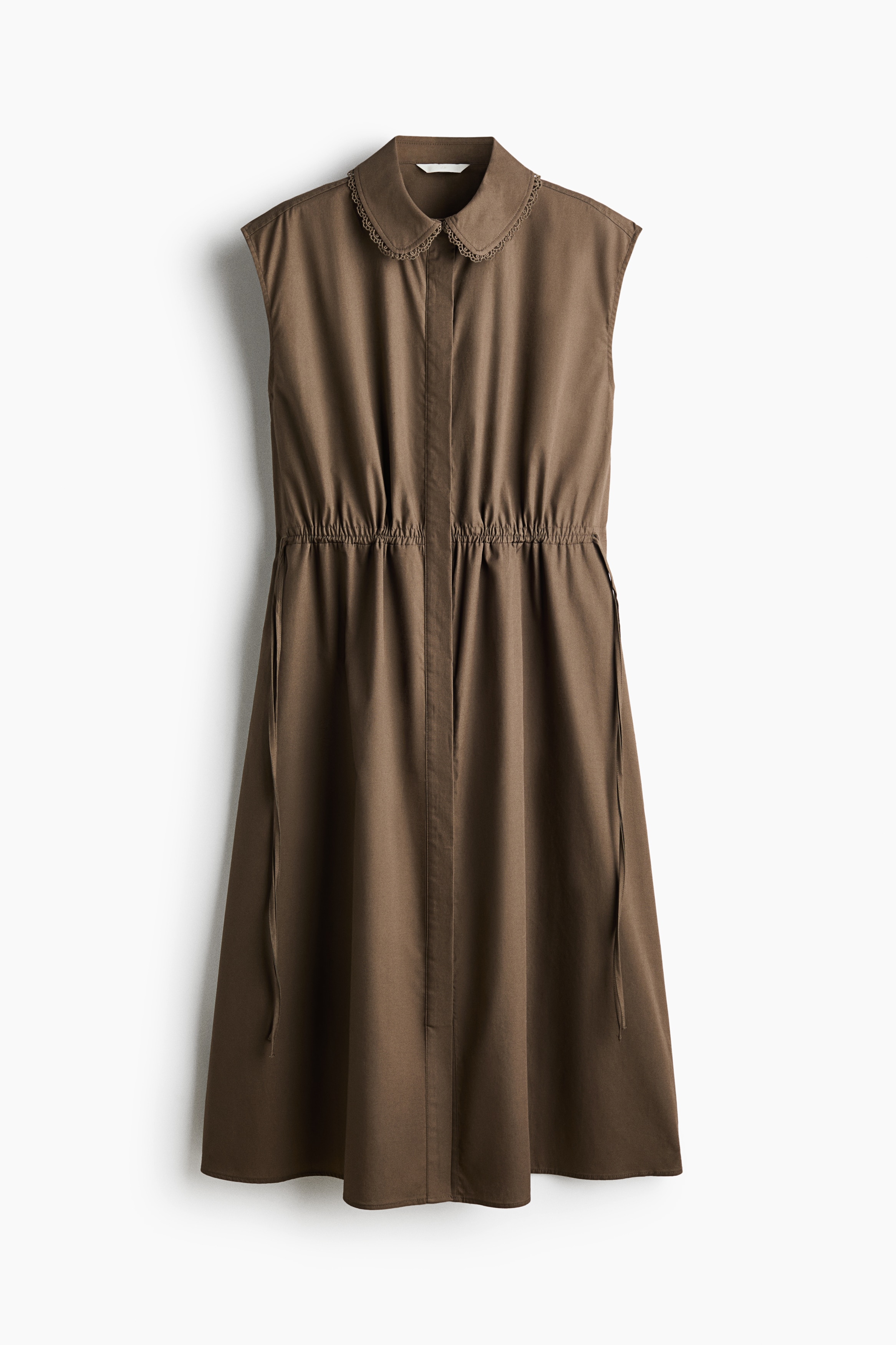 The model is wearing a calf-length, sleeveless brown cotton dress. The dress features a lace-trimmed collar, a concealed button placket, a gathered drawstring waist, and a gently flared skirt, paired with black pointed-toe flat shoes.