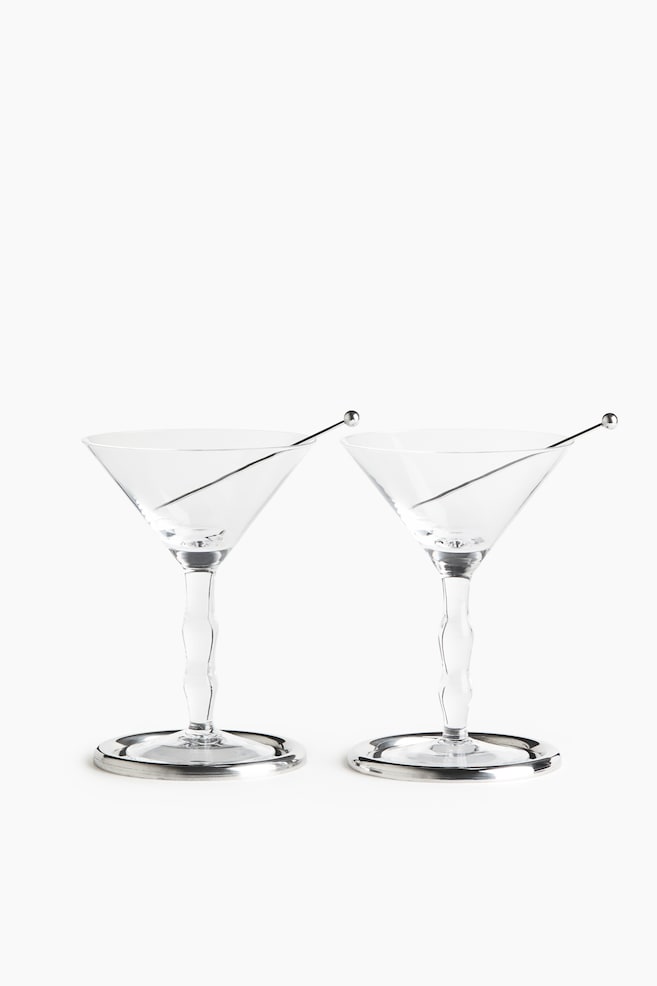 Une paire de verres Ã  martini en verre transparent est disposÃ©e cÃ´te Ã  cÃ´te, tournÃ©e vers lâavant, avec des coupes coniques, des pieds ondulÃ©s sculptÃ©s et des bases circulaires de ton argentÃ©. Chaque verre contient un fin pic Ã  cocktail en mÃ©tal argentÃ© avec une petite extrÃ©mitÃ© sphÃ©rique, posÃ© en diagonale Ã  lâintÃ©rieur de la coupe. - 1