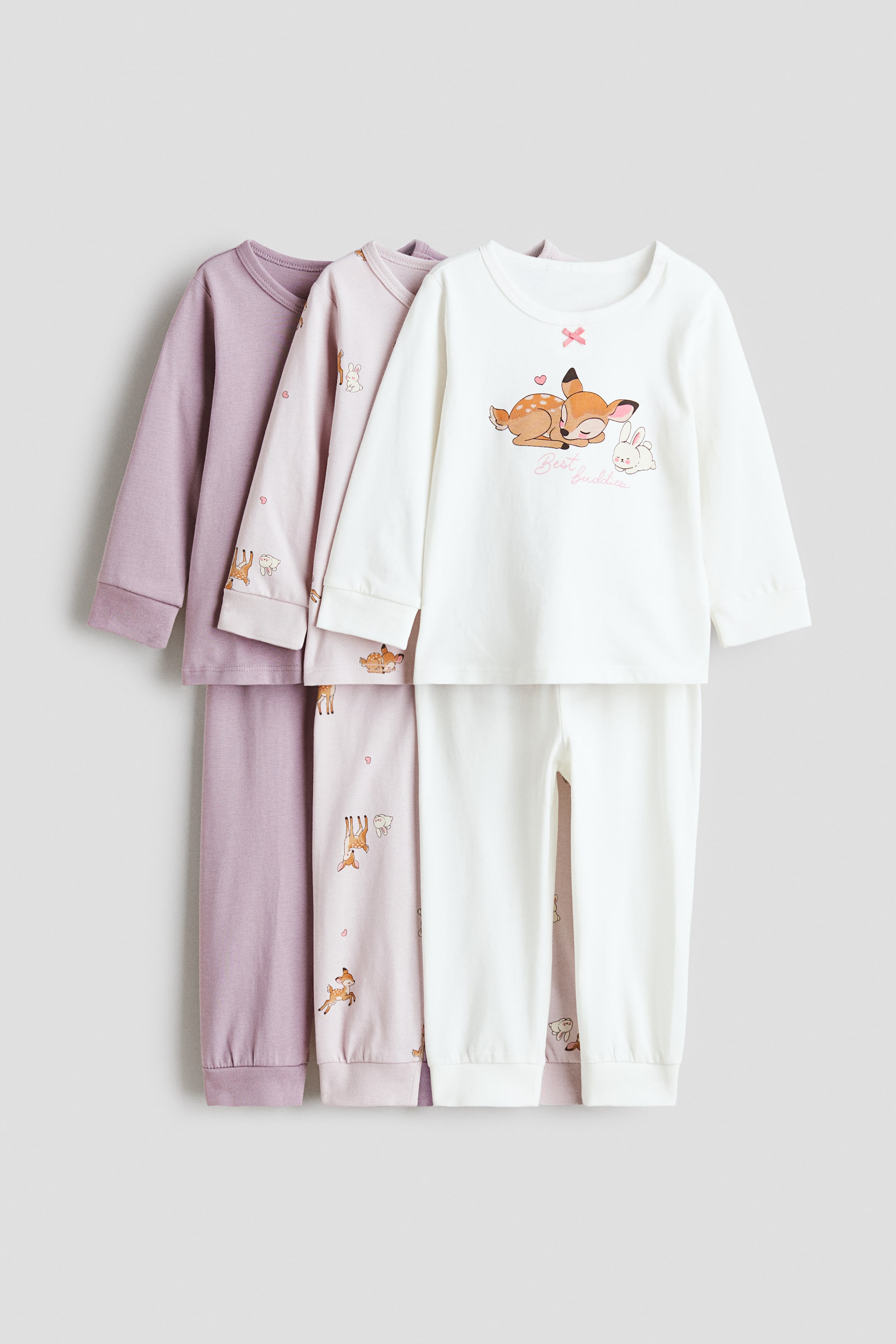 Zobrazit větší obrázek: Sada tří bavlněných pyžam zobrazuje tři soupravy s dlouhým rukávem a kalhotami, naskládané na sobě tak, aby byly vidět jejich přední strany. Přední souprava v odstínu slonové kosti má na horním dílu potisk spící srnky a králíčka, zatímco prostřední světle růžová souprava je vzorovaná srnkami, králíčky a srdíčky. Zadní světle fialová souprava je v jednobarevném provedení a všechny kalhoty mají elastické manžety u kotníků.