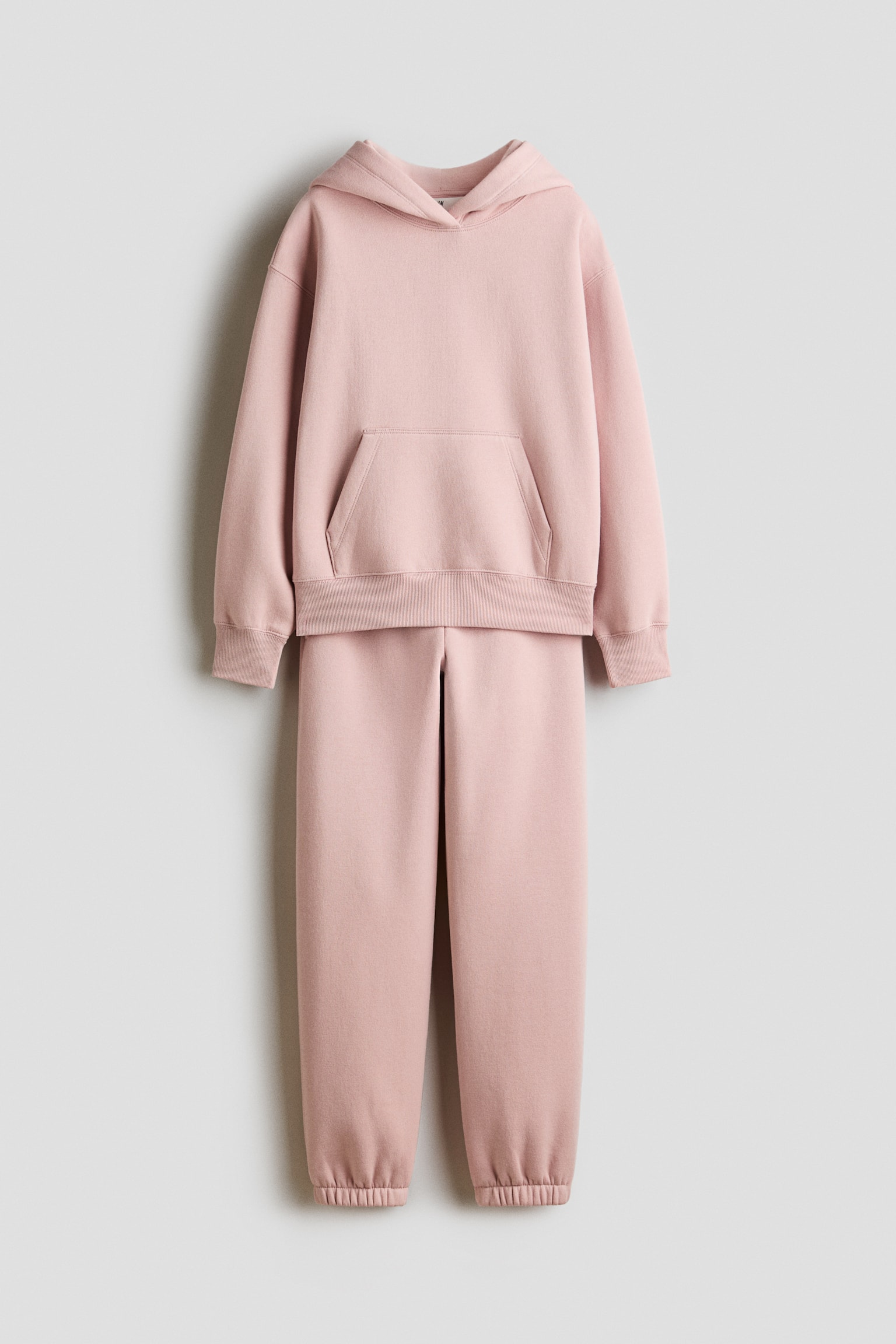 Se muestra de frente un conjunto deportivo de dos piezas en rosa claro, compuesto por una sudadera con capucha y pantalones de sudadera a juego. La sudadera presenta un bolsillo tipo canguro frontal con puños y dobladillo acanalados, mientras que los pantalones terminan con puños elásticos en los tobillos.