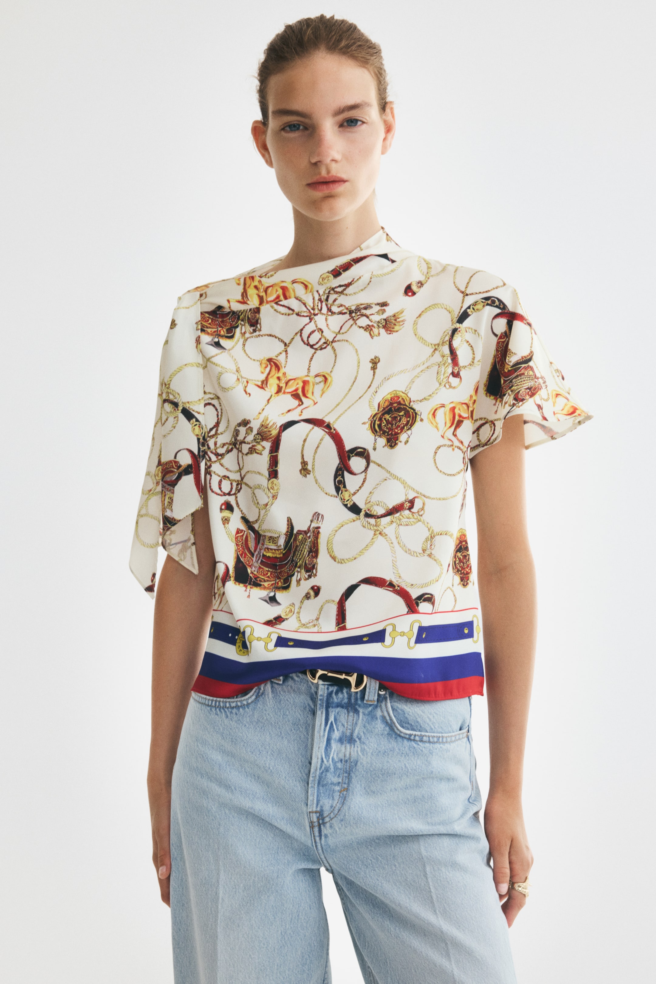 Ver imagem maior: Um top em sarja acetinada com padrão de inspiração equestre é usado pela modelo, combinado com jeans azuis de lavagem clara. O top apresenta um decote assimétrico com um detalhe drapeado sobreposto de um lado e uma manga curta estilo batwing do outro.