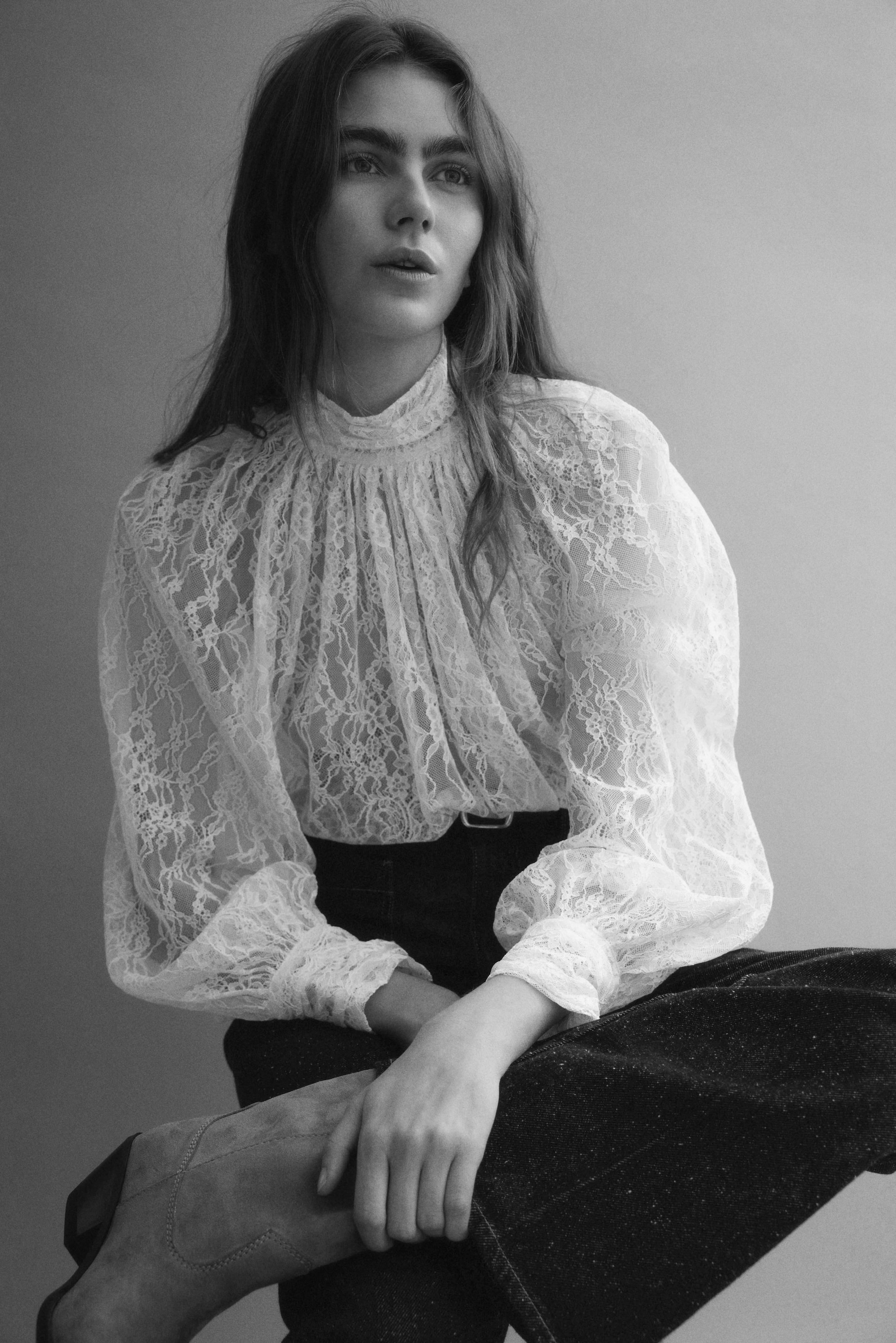 View larger image: The white lace blouse features a high, gathered mock neck and voluminous long sleeves that gather into buttoned cuffs. The sheer blouse showcases intricate floral lace patterns and is worn tucked into dark pants, revealing its full silhouette.