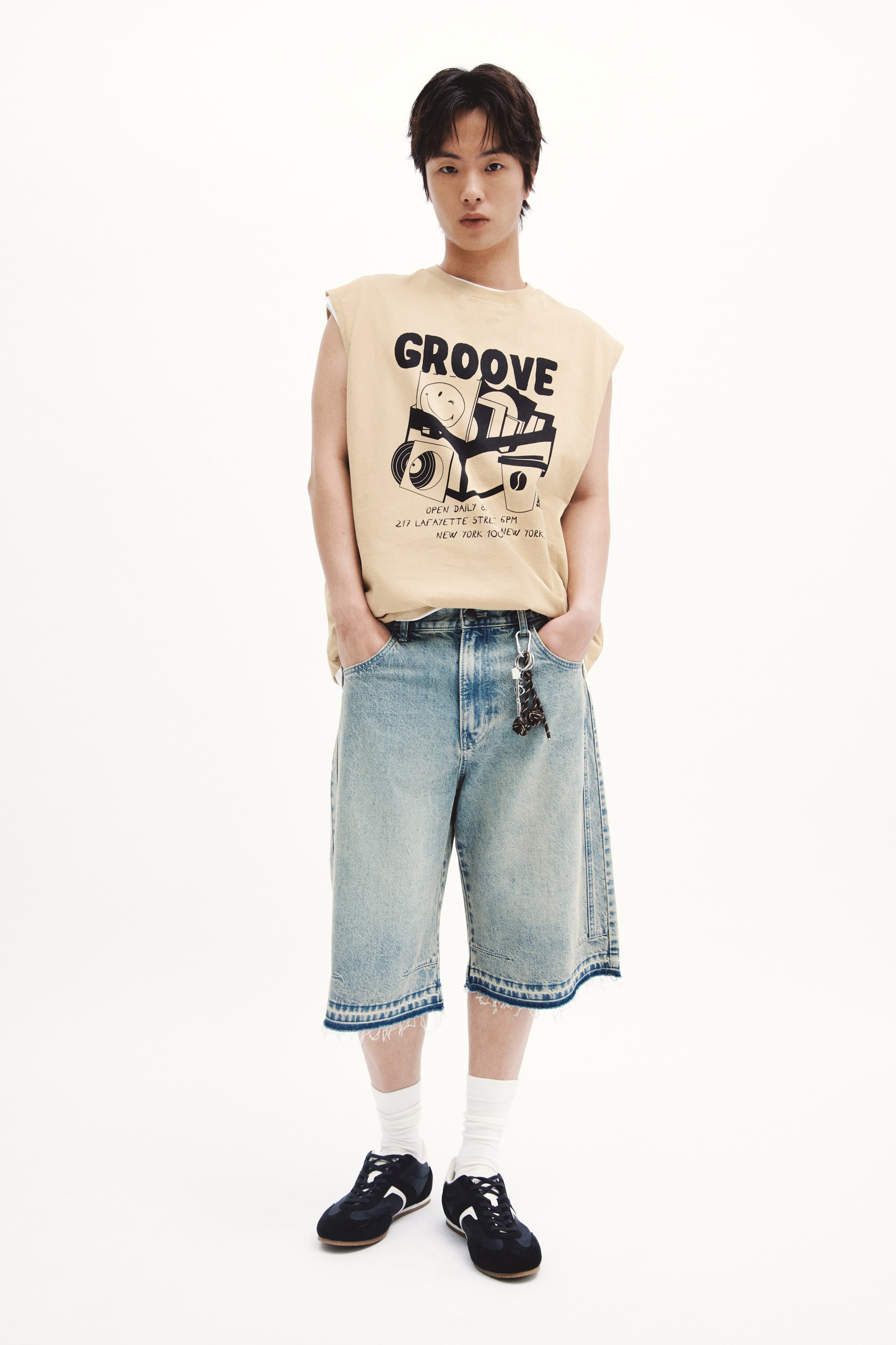 拡大画像を表示: このコーディネートは、カメラ、カセットテープ、コーヒーカップをあしらった「GROOVE」グラフィックプリントが施された、ライトタンのノースリーブTシャツで構成されています。これに合わせるのは、ラフでリラックスしたフィット感のあるフェードライトブルーデニムショーツで、裾は切りっぱなしのフリンジデザインになっています。ベルトループにはキーチェーンが掛けられています。ホワイトのクルーソックスと、ホワイトアクセントが施されたダークスニーカーがこのコーディネートを完成させます。