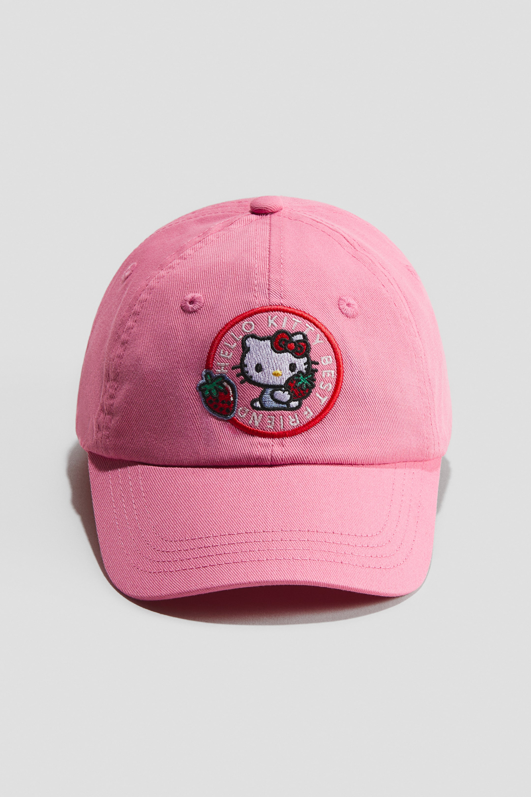 View larger image: A pink cap in soft cotton twill is facing front, featuring a circular embroidered patch. The patch showcases Hello Kitty holding strawberries, bordered by the text "HELLO KITTY BEST FRIENDS."