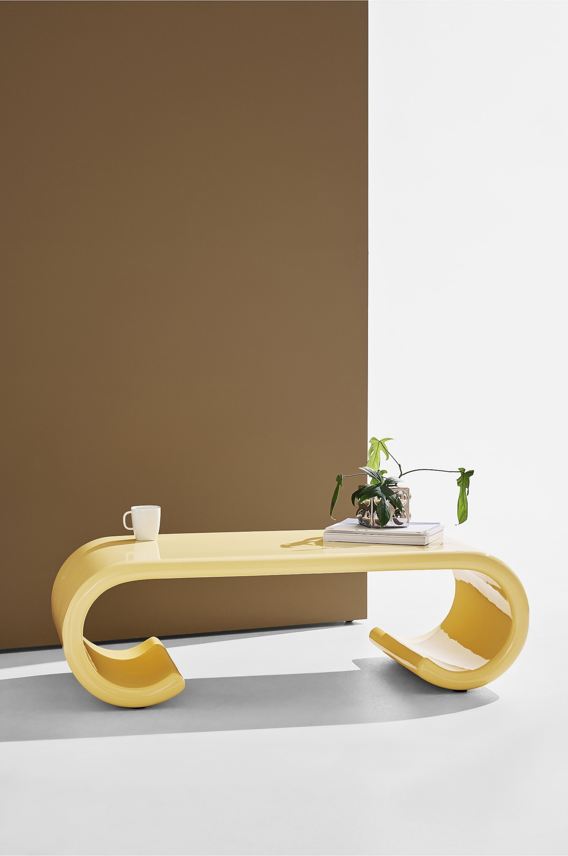 Agrandir l'image: Une table basse brillante jaune vif présente un design continu et incurvé, formant une surface supérieure plate et deux grands pieds en forme de U. Une tasse blanche, une pile de livres et une petite plante en pot reposent sur son dessus.