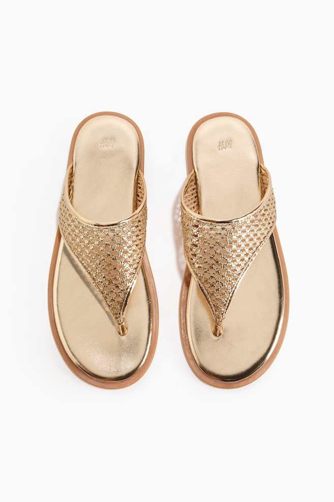 Um par de sandálias douradas de dedo apresenta tiras metálicas entrelaçadas e palmilhas douradas lisas, apresentadas voltadas para a frente. Cada sandália exibe uma sola castanho-clara e um logótipo H&M na sua palmilha. - 1