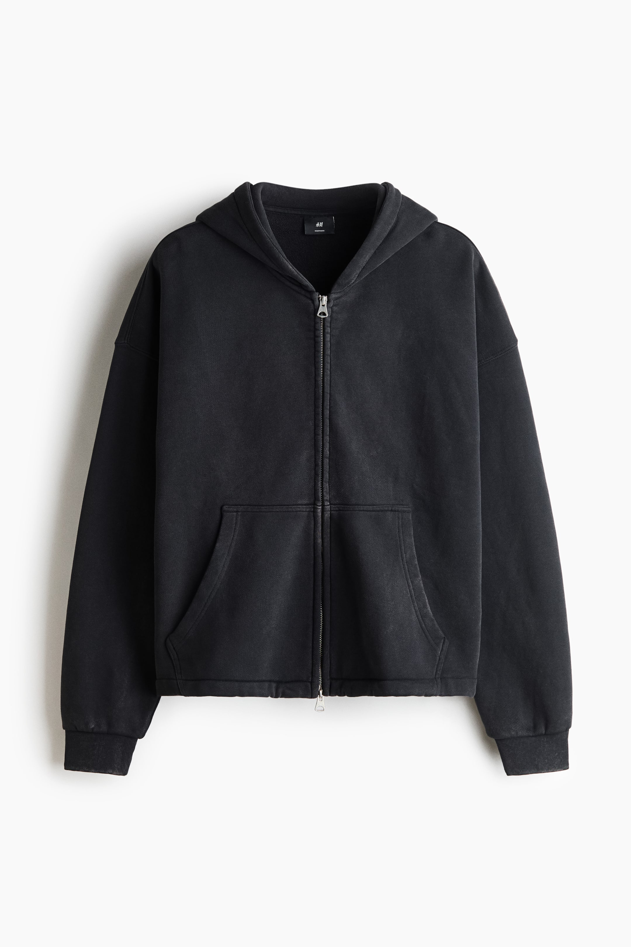 Xem ảnh lớn hơn: Chiếc áo hoodie zip màu đen washed, kiểu dáng boxy này được hiển thị ở góc nhìn chính diện, với mũ trùm lót jersey, vai trễ và tay dài bo gấu rib. Sản phẩm nổi bật với khóa kéo hai chiều phía trước và túi kangaroo chẻ đôi, tôn lên phom dáng oversized, rộng thùng thình.
