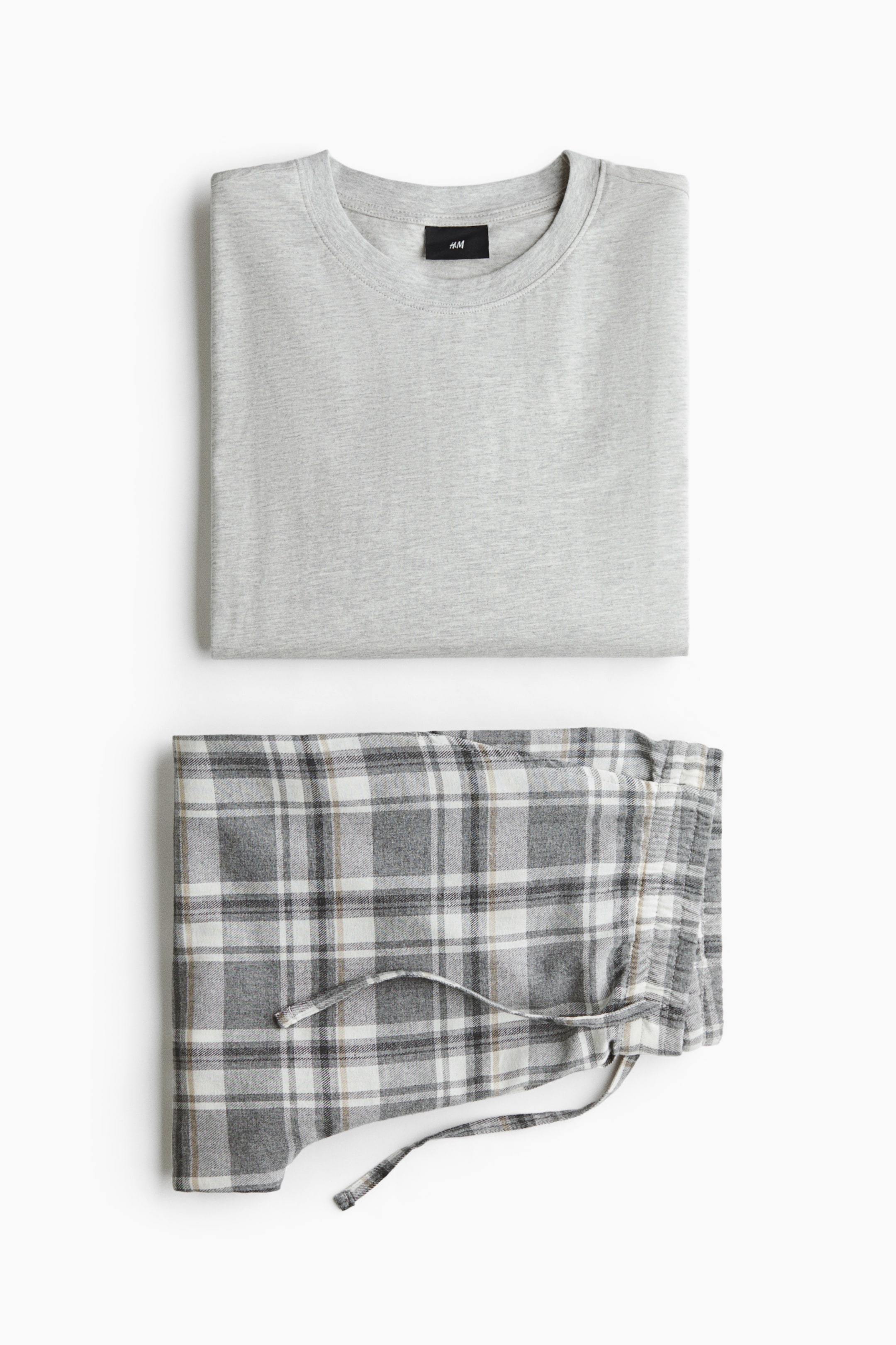 Se større billede: Et pyjamassæt vises foldet fladt og består af en lysegrå top i bomuldsjersey med rund halsudskæring samt et par flannelbukser i grå, hvid og beige tern med synlig snøre i taljen.