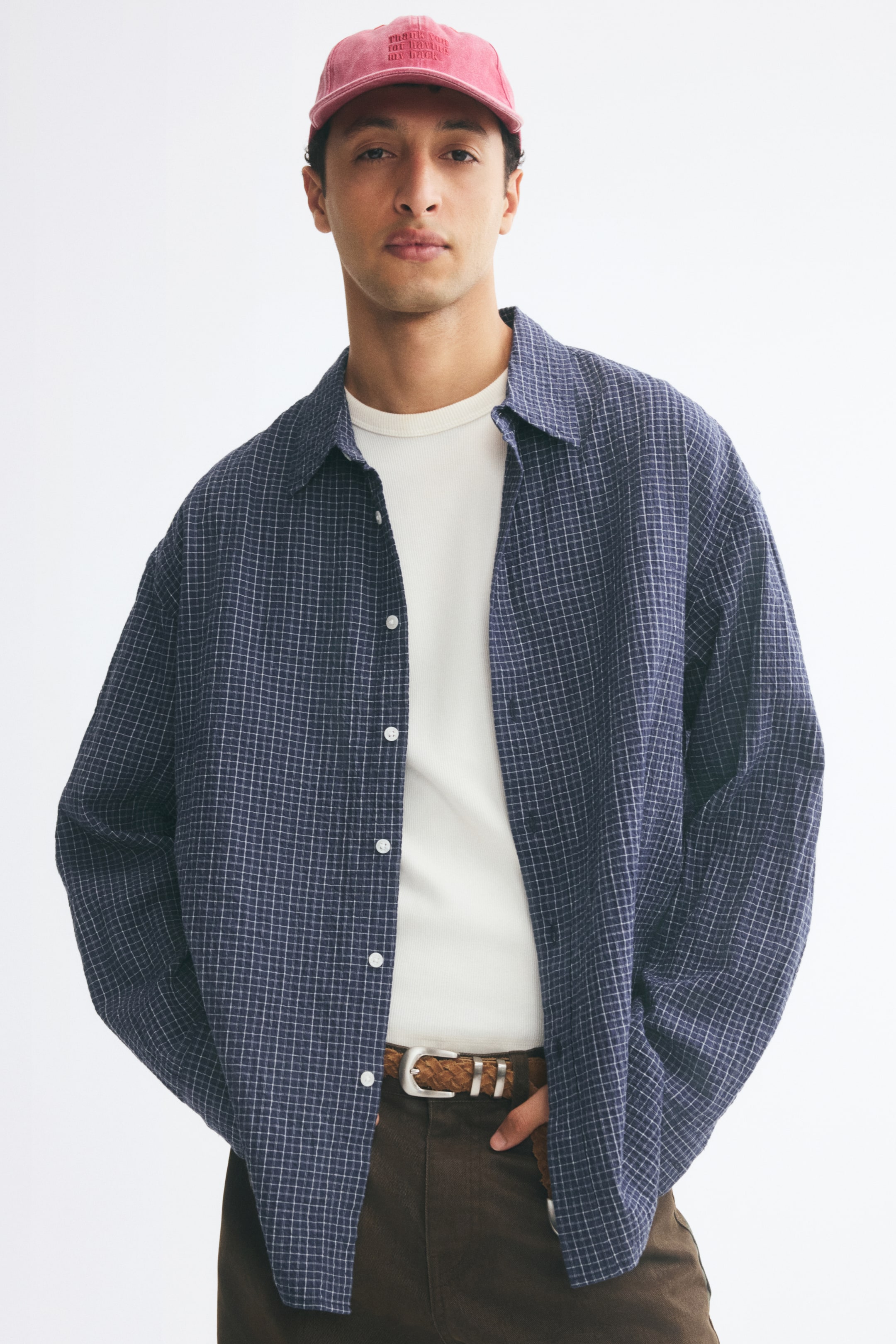 View larger image: A relaxed-fit, long-sleeved shirt with a blue-and-white grid pattern and a turn-down collar is worn open by the model. The shirt is layered over a white T-shirt and paired with brown trousers.