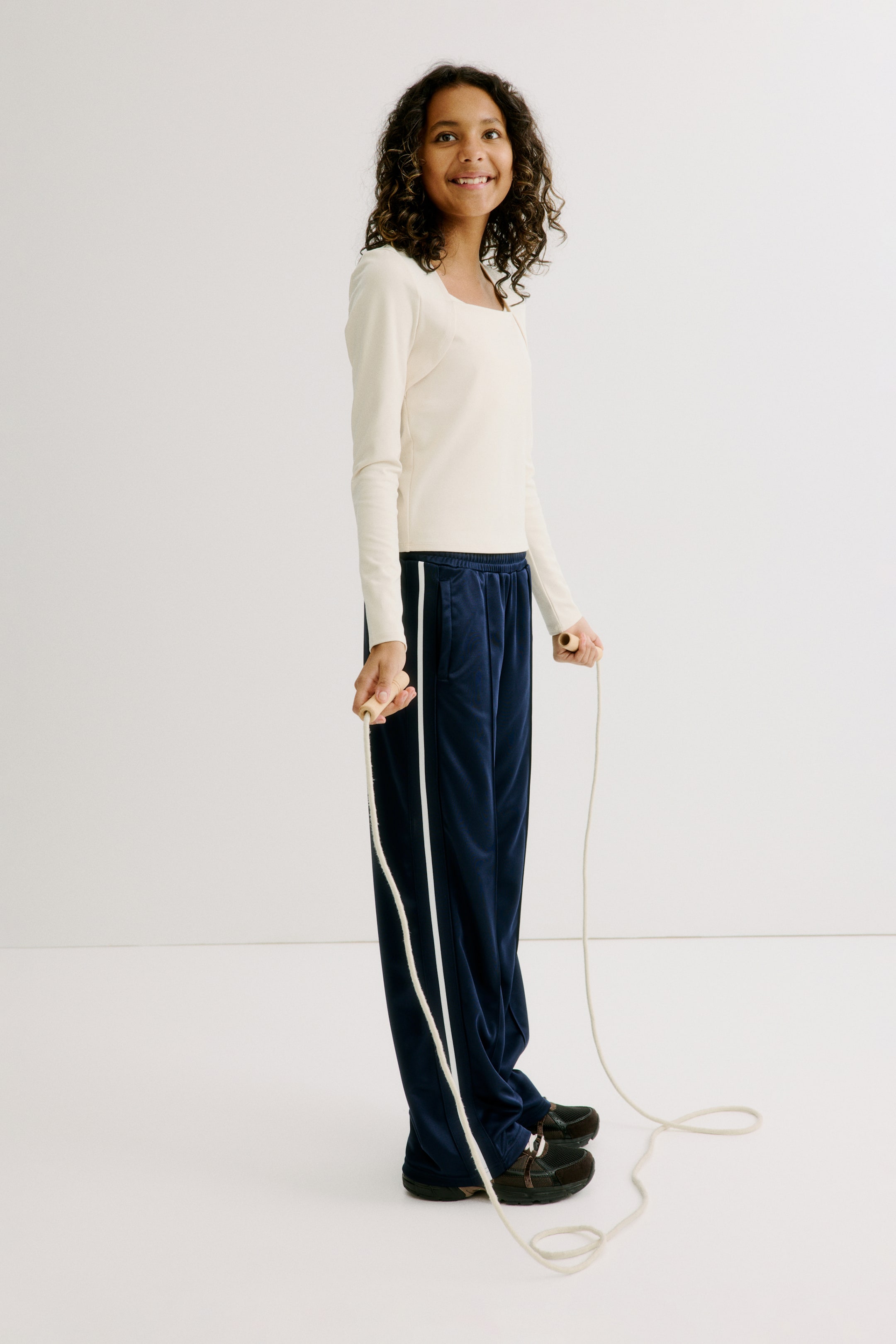 Ampliar la imagen: Un top de manga larga color crema con un escote cuadrado está siendo utilizado, presentando una silueta ajustada. Está combinado con un pantalón de chándal de terciopelo azul marino, que presenta una franja blanca que corre a lo largo de cada costura lateral y una pretina elástica, mostrando un ajuste holgado y de pierna ancha.