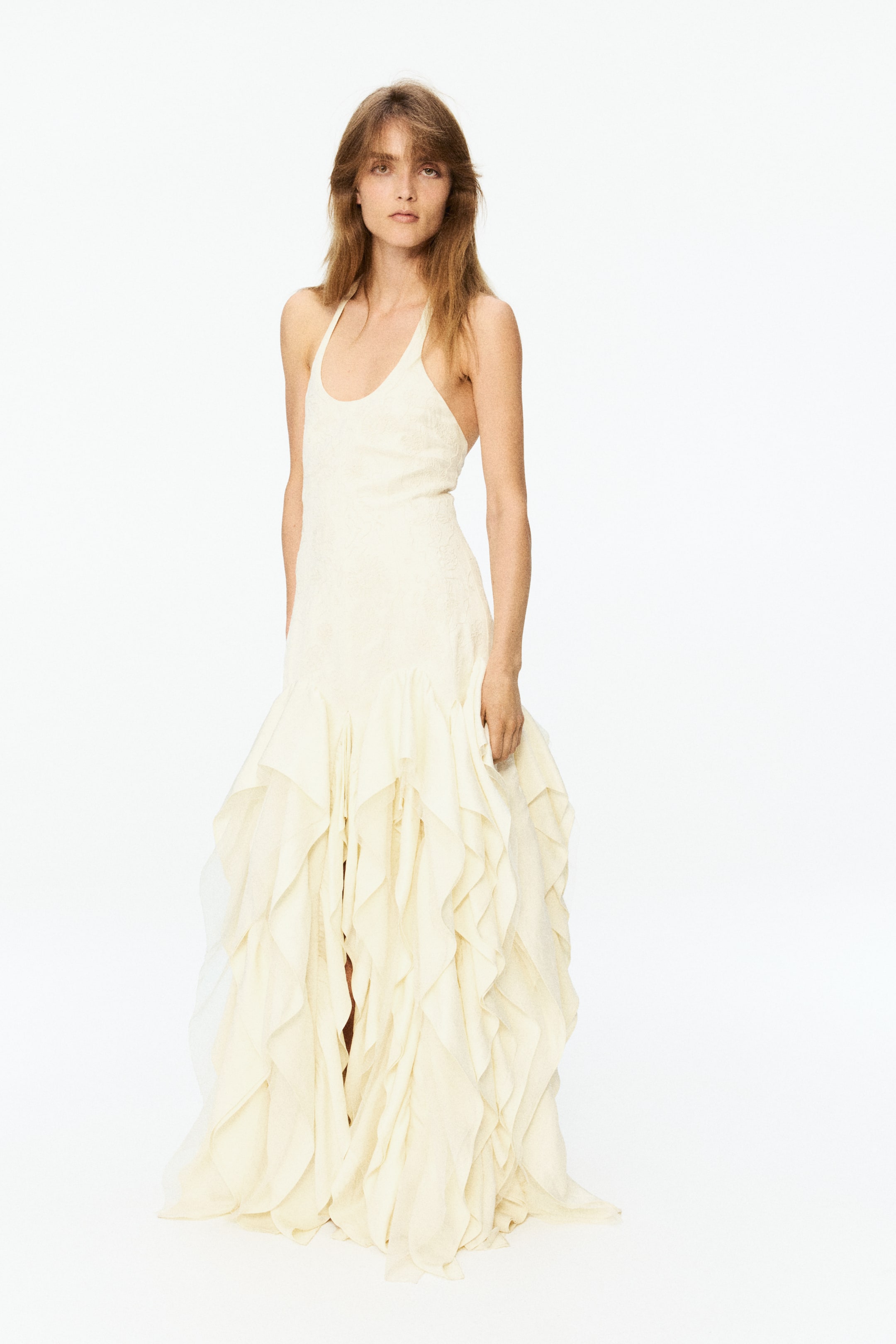 View larger image: The cream-colored halter-neck dress showcases a fitted bodice with a subtle embossed pattern. Its full-length skirt flows with numerous layered ruffles, starting from mid-thigh and extending to the floor, creating a voluminous silhouette.