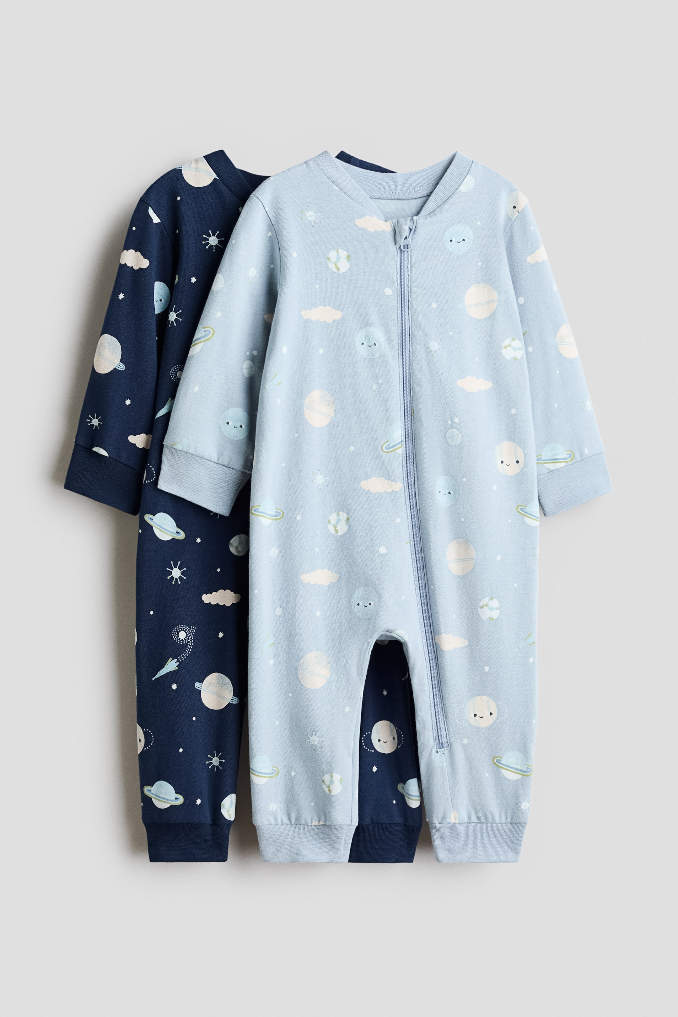 Ver imagem maior: Dois babygrows são apresentados, com o azul claro voltado para a frente e exibindo um fecho de correr de comprimento total. Ambos os babygrows têm mangas compridas, pernas compridas com punhos canelados e uma estampa temática espacial de planetas, estrelas e corpos celestes sorridentes; o segundo babygrow é azul escuro e está parcialmente visível por trás do primeiro.