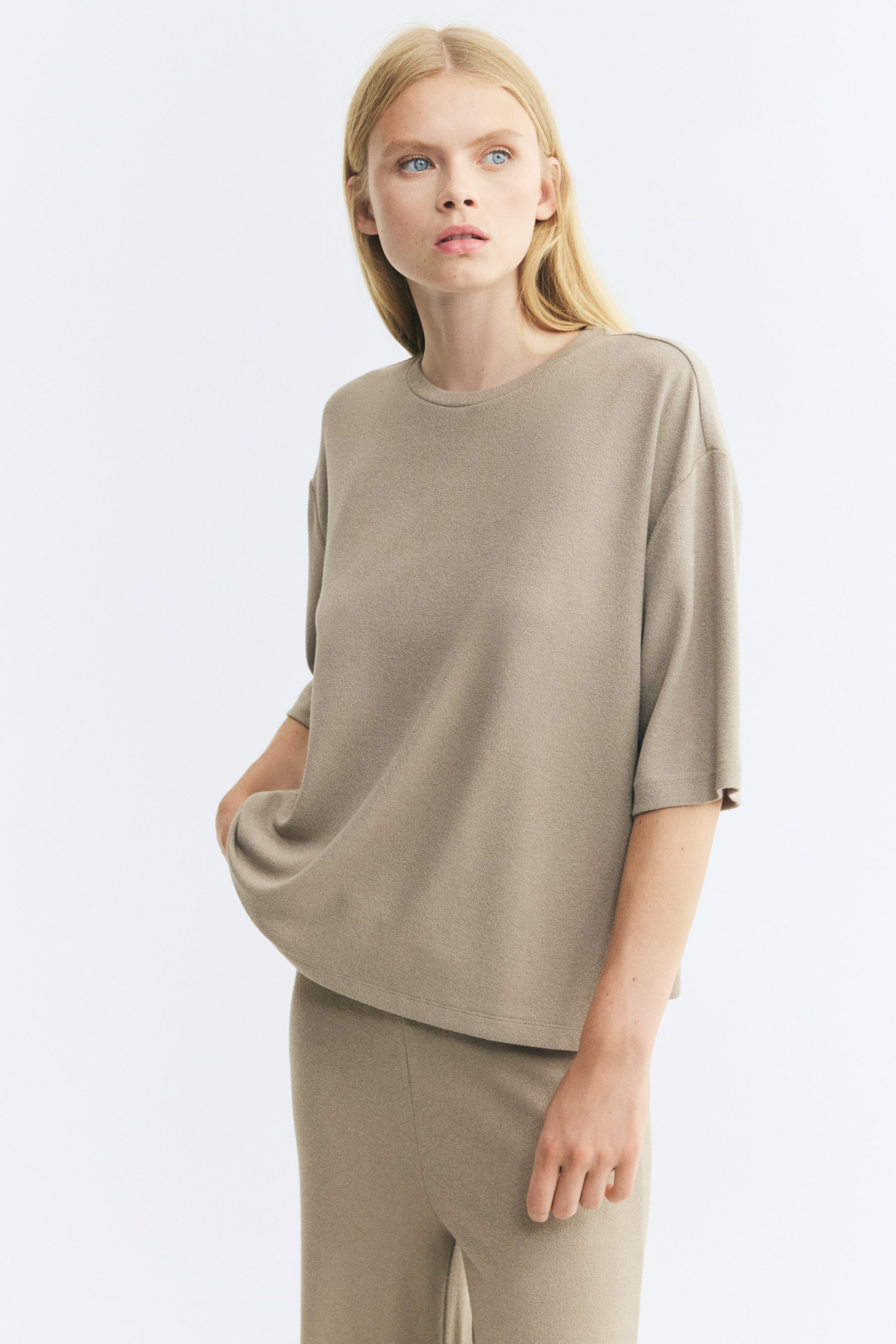 View larger image: The model is wearing an oversized beige top featuring a round neckline, dropped shoulders, and 3/4-length sleeves. The top is styled with matching trousers, and the model is resting one hand in a pocket, creating a soft drape at the hem.