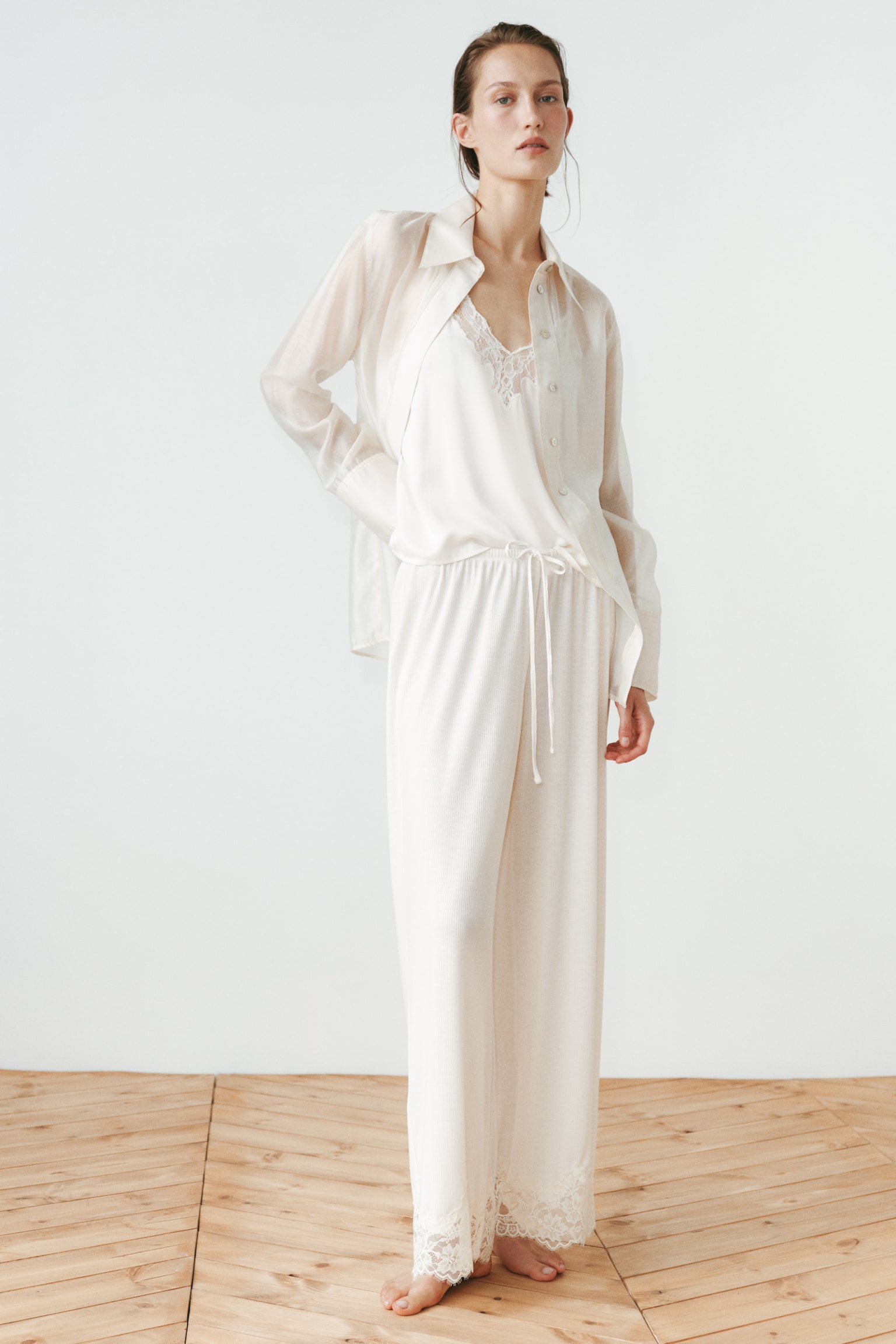 Cream-colored ribbed pants are hanging front-facing, featuring an elasticated waistband with a central drawstring tie and wide lace trim adorning the bottom hems of both legs. - 1