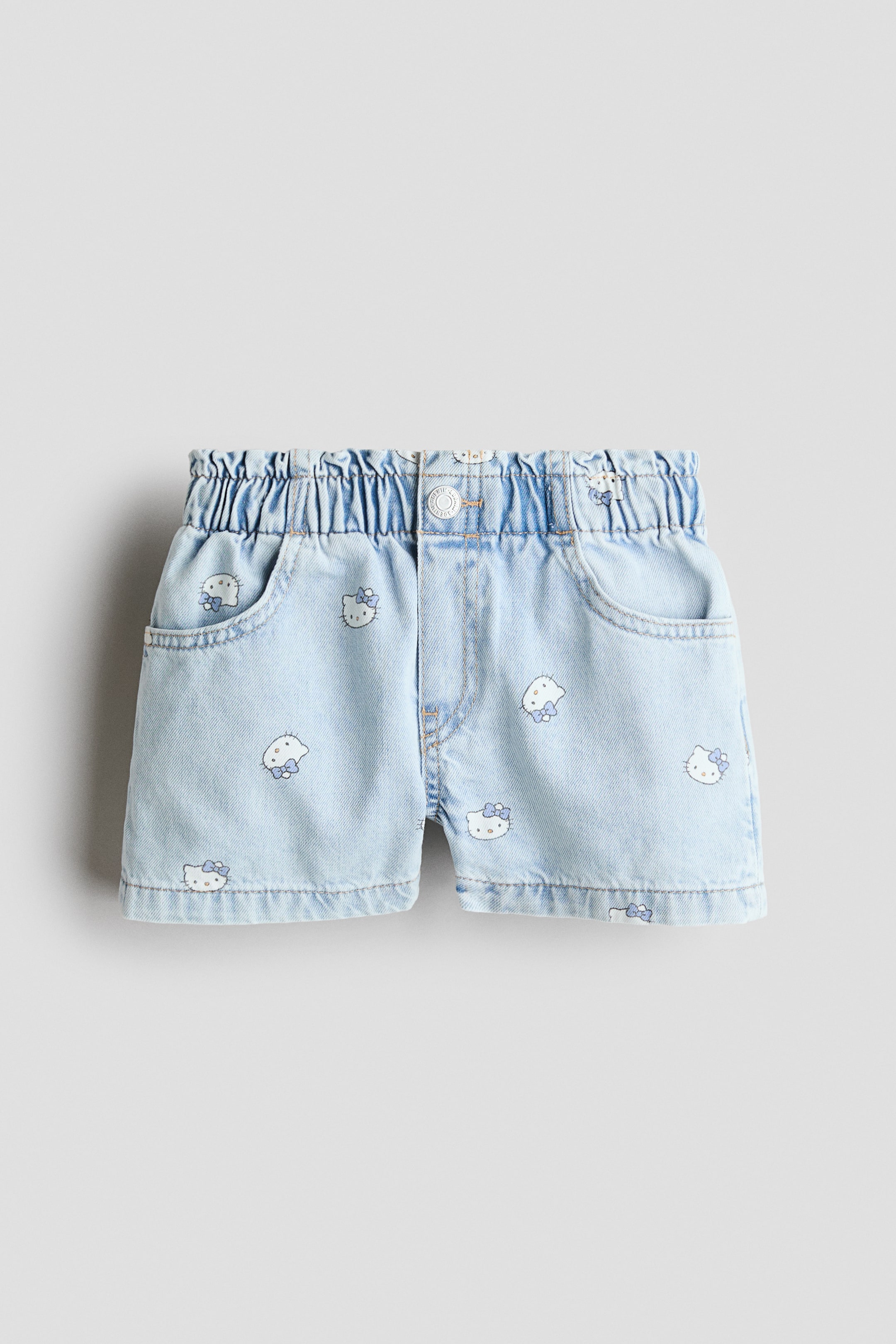 View larger image: The front of these light blue cotton denim shorts showcases a relaxed fit and an all-over print of white cartoon character heads adorned with blue bows. They are crafted with an elasticated paper bag waist, a mock fly with a button, and diagonal front pockets.