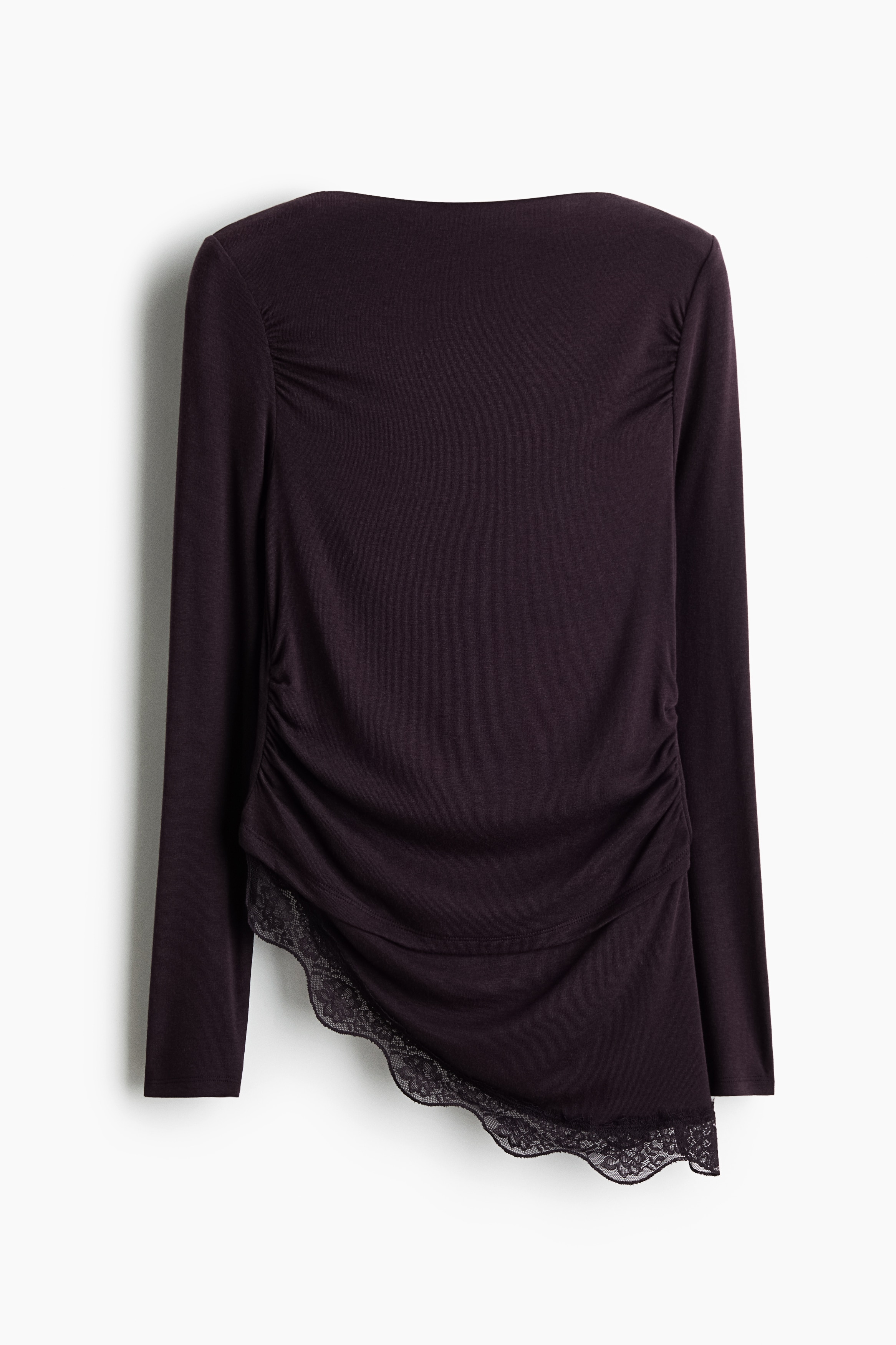 A dark plum, fine-ribbed jersey top is being worn by the model, featuring a boat neckline, long sleeves, draped gathers, and an asymmetrical hem with scalloped lace trim. The model is lightly holding the gathered fabric near the waist, styling the top with dark denim trousers.