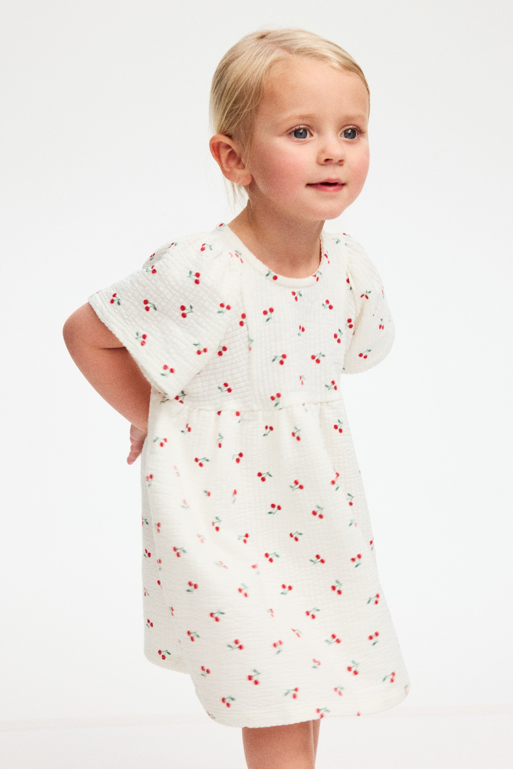 View larger image: The child's off-white dress features a crinkled texture, patterned with small red cherries and green stems. It has a round neck, short puffed sleeves and a gathered empire waistline, creating a flowing skirt as it faces forward, angled slightly to the right.