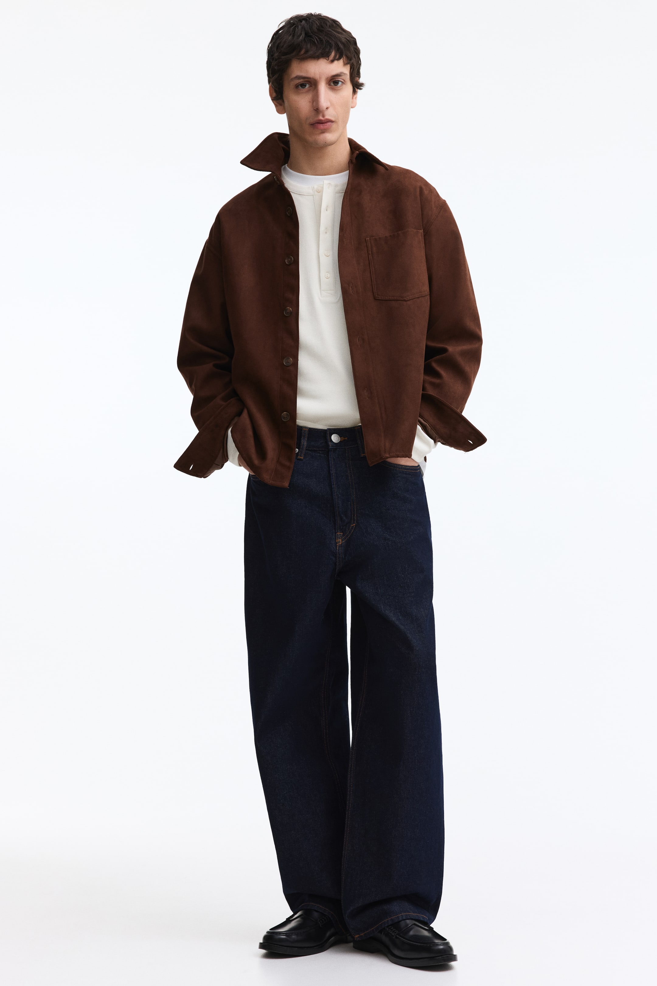 Ingrandisci l'immagine: Una sovracamicia marrone con una texture simile al camoscio è indossata aperta su una maglia henley bianca, caratterizzata da un colletto a punta, bottoni scuri lungo l'abbottonatura, maniche lunghe con polsini sbottonati e una tasca applicata sul petto sinistro.