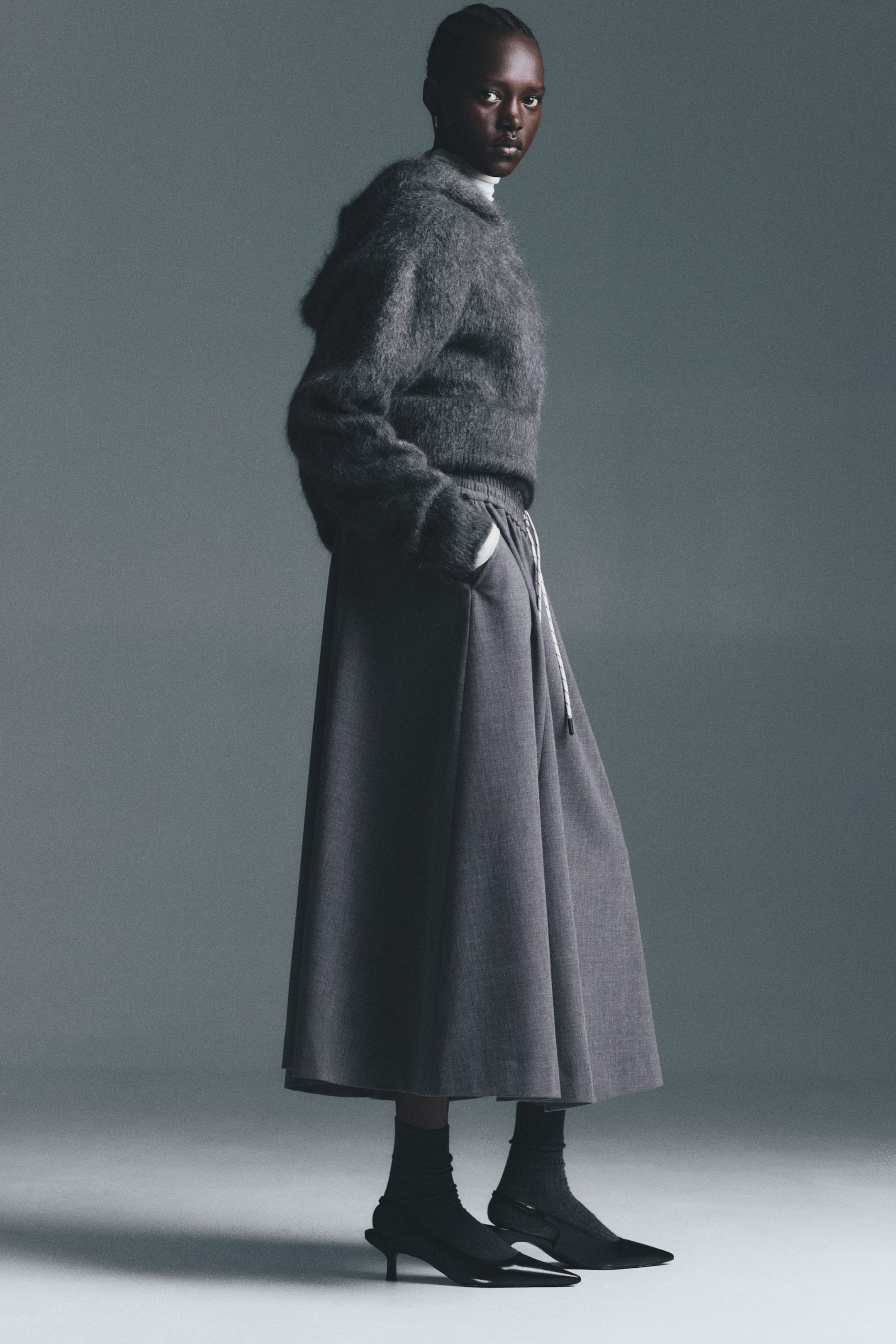 View larger image: A dark gray fuzzy hooded sweater is layered over a white turtleneck, paired with a dark gray midi-length skirt featuring an elastic waistband and a white drawstring. The outfit is completed with black pointed-toe kitten heels and black ribbed socks, presented from a slight side angle.