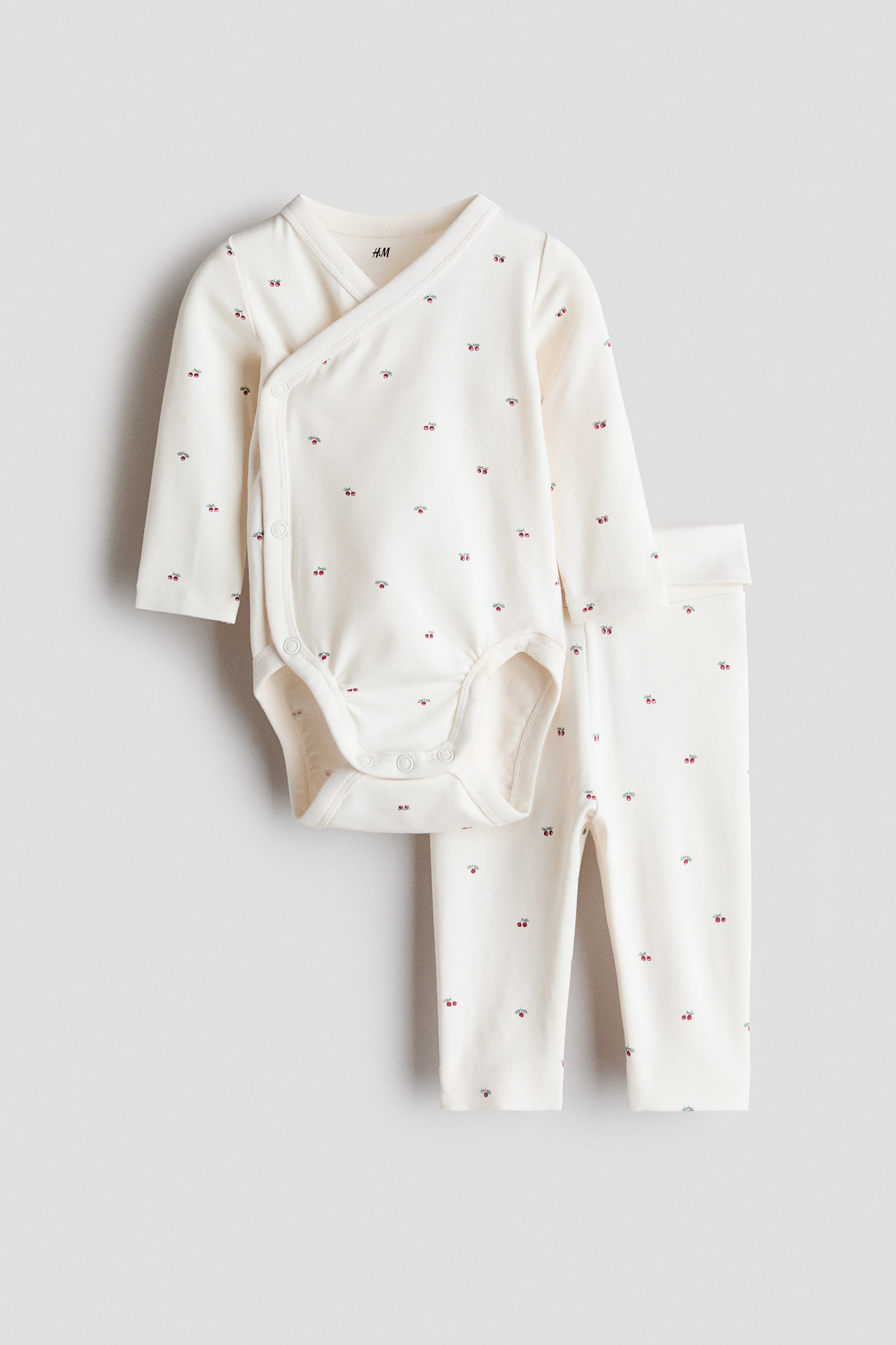 View larger image: A cream-coloured baby clothing set showcases a wrapover bodysuit and coordinating leggings, both adorned with a small red cherry pattern. The bodysuit features a V-neck, long sleeves, and press-stud closures along the side and at the crotch, with gathered leg openings for a snug fit. The matching leggings display a foldover waist.