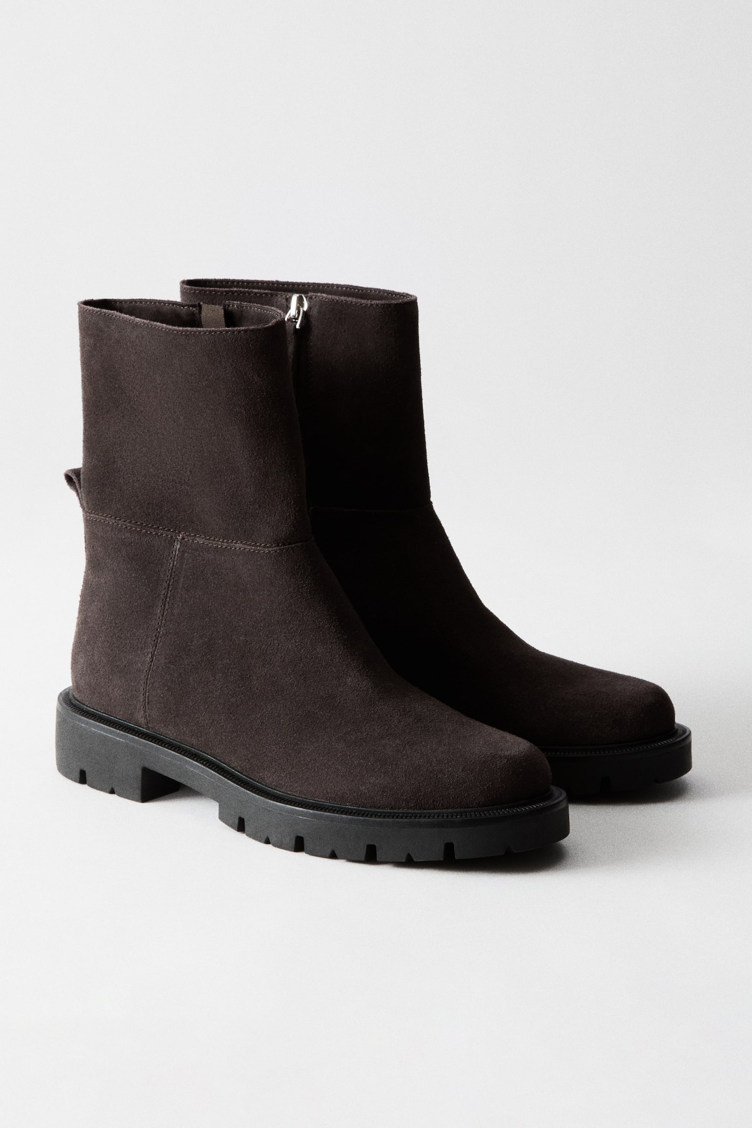 A dark brown suede ankle boot is facing right, featuring a mid-height shaft with visible stitching details and a small pull-tab at the back. It rests on a thick black lugged sole with a block heel. - 1