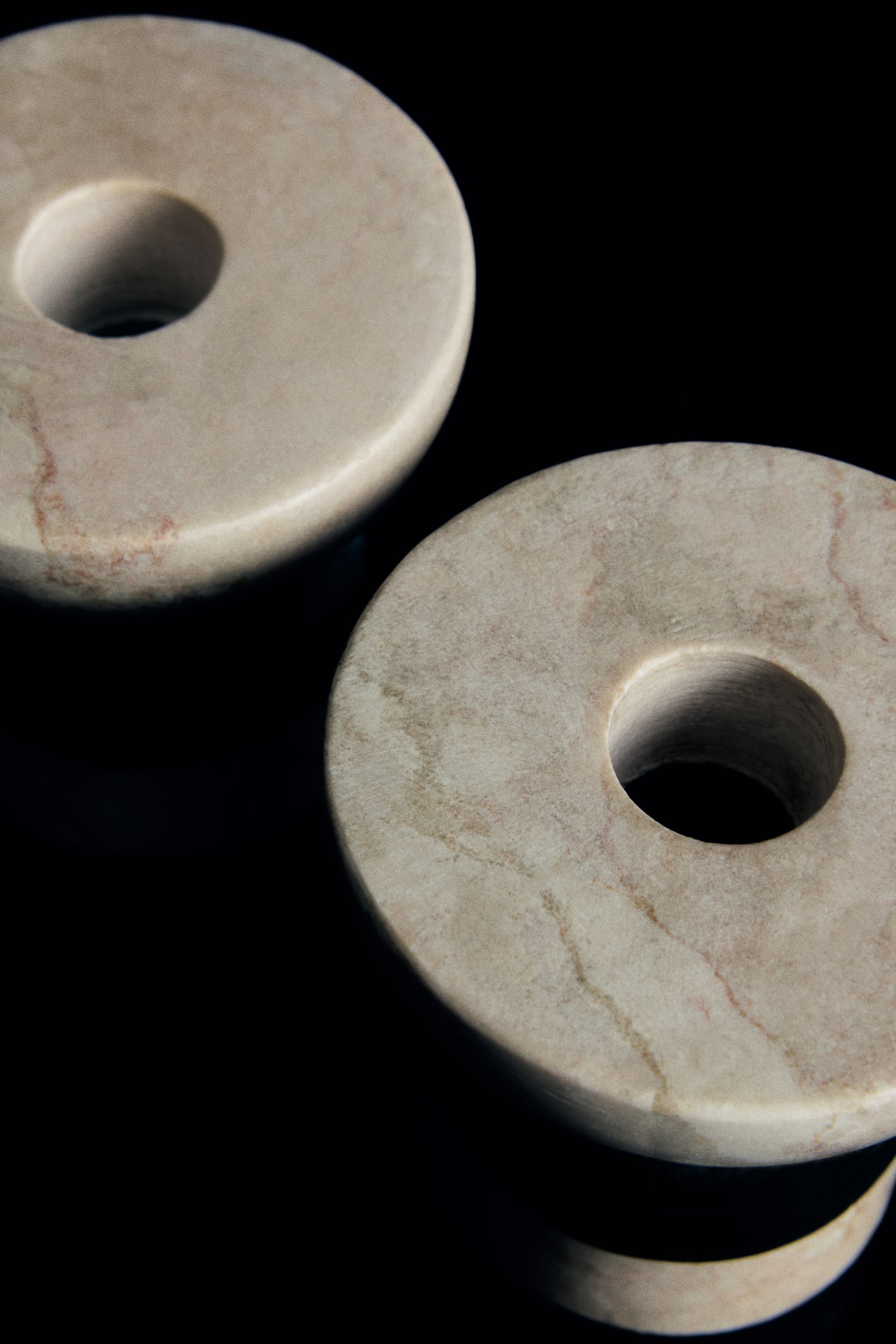 Ampliar la imagen: Dos candelabros pequeños de mármol muestran sus copas circulares acampanadas desde un ángulo elevado, resaltando las vetas naturales beige claro y marrón del mármol. El candelabro del primer plano revela una banda oscura en contraste alrededor de su cuerpo cilíndrico, debajo de la parte superior acampanada.