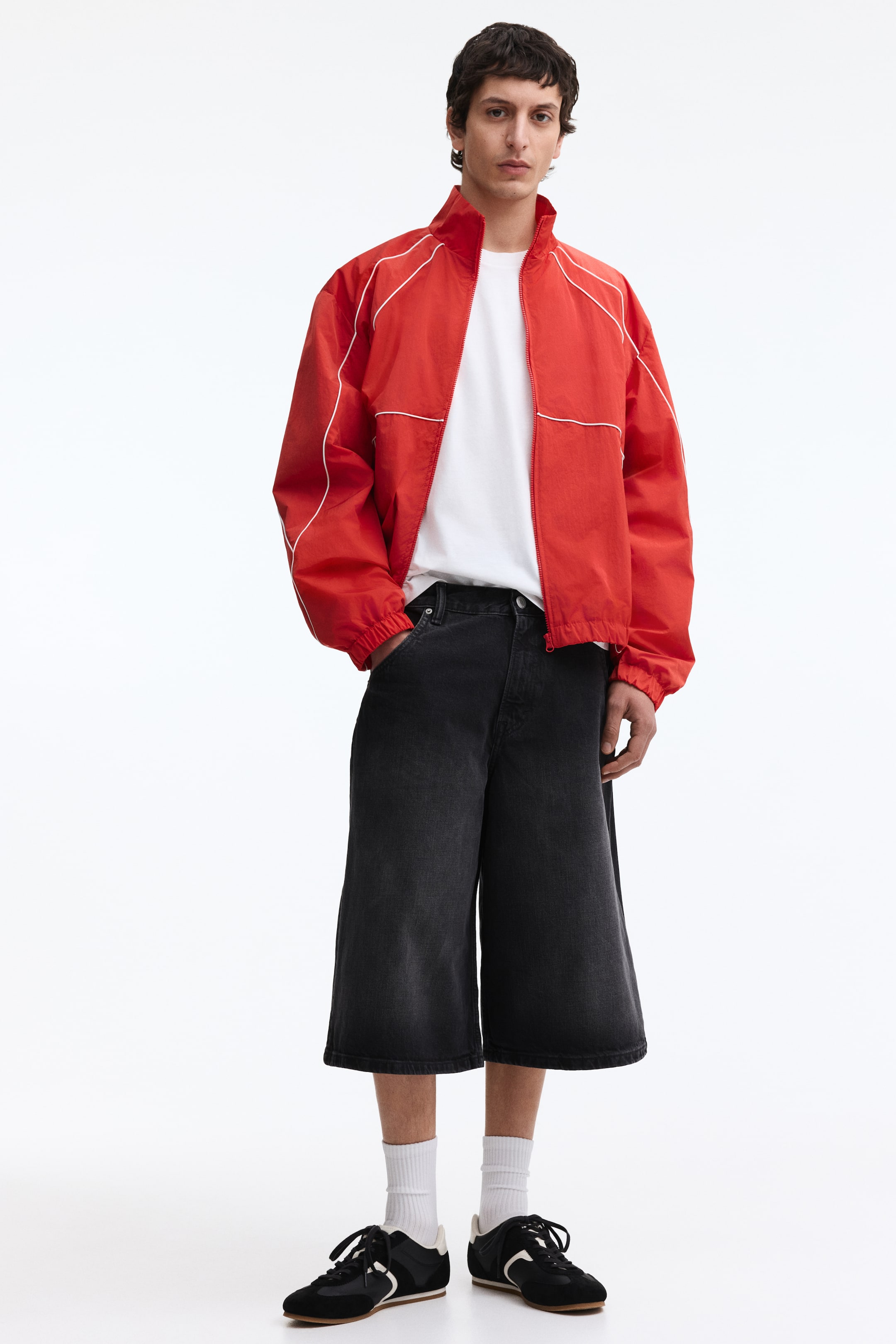 Voir une image plus grande: Une veste de survêtement rouge à fermeture éclair complète, avec un col montant et des passepoils blancs sur les épaules et la poitrine, est portée par-dessus un t-shirt blanc à col rond, associée à un pantalon en denim noir délavé à jambes larges se terminant au-dessus des chevilles, et des baskets noires avec des semelles, lacets et bandes latérales blancs, accompagnées de chaussettes blanches côtelées.