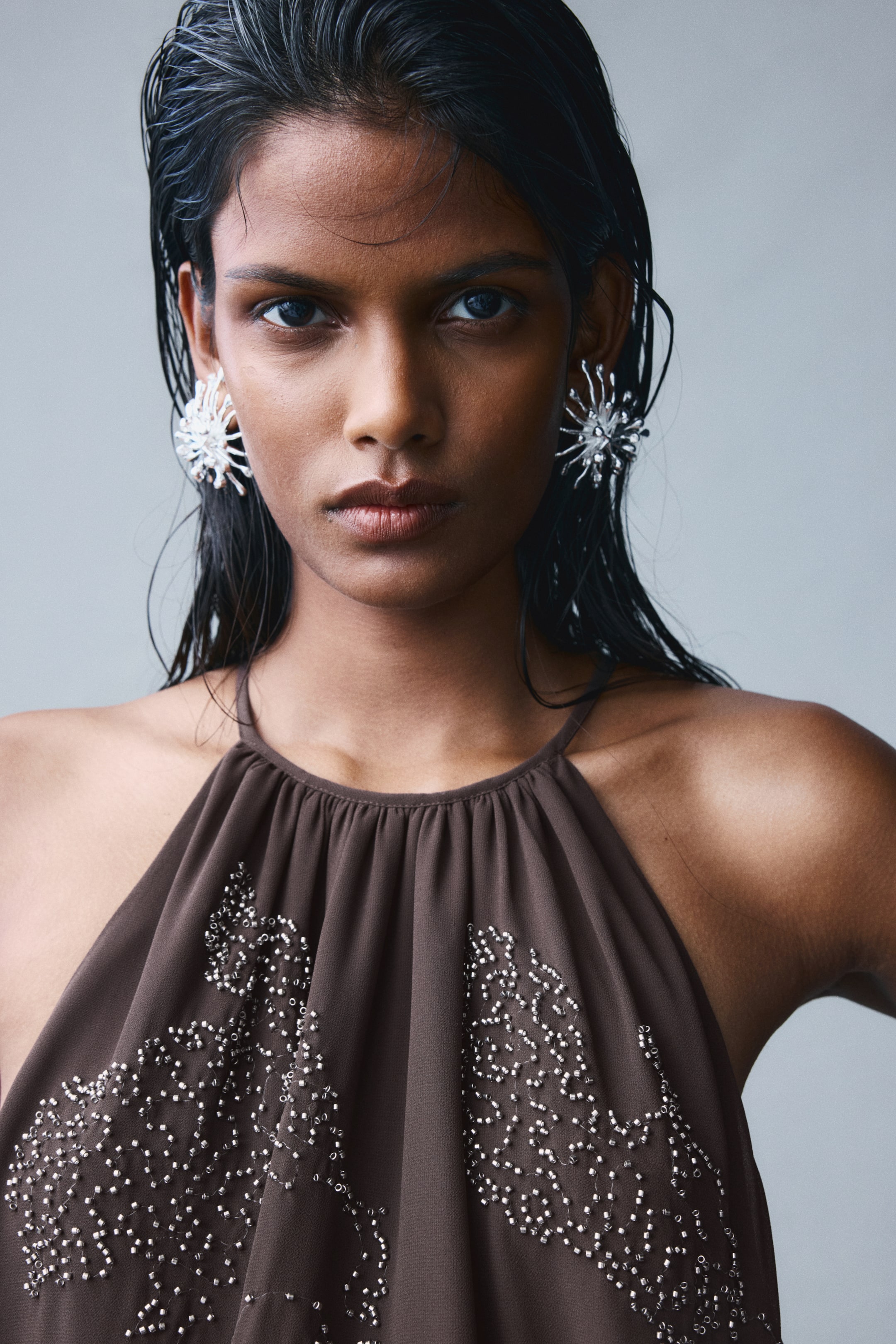 View larger image: A brown chiffon top featuring a gathered neckline, thin shoulder straps, and intricate silver beaded designs on the front is being worn by the model. The top is styled with large, textured silver earrings that coordinate with the beadwork.