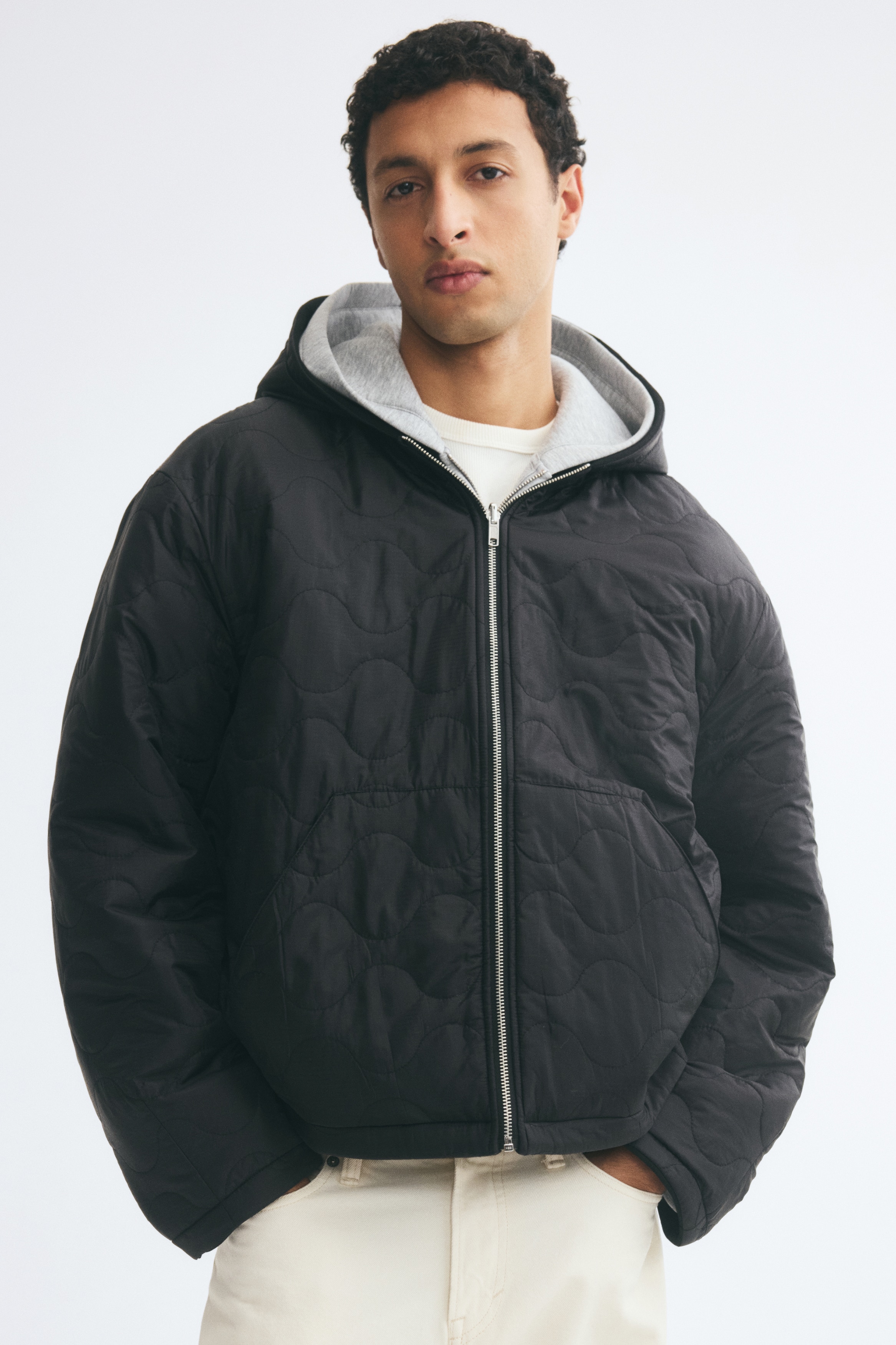 Una giacca nera con cappuccio Ã¨ mostrata frontalmente, caratterizzata da una trama ripstop trapuntata con motivo a cipolla. Il design include una zip centrale color argento, spalle scese e tasche frontali a filo oblique.