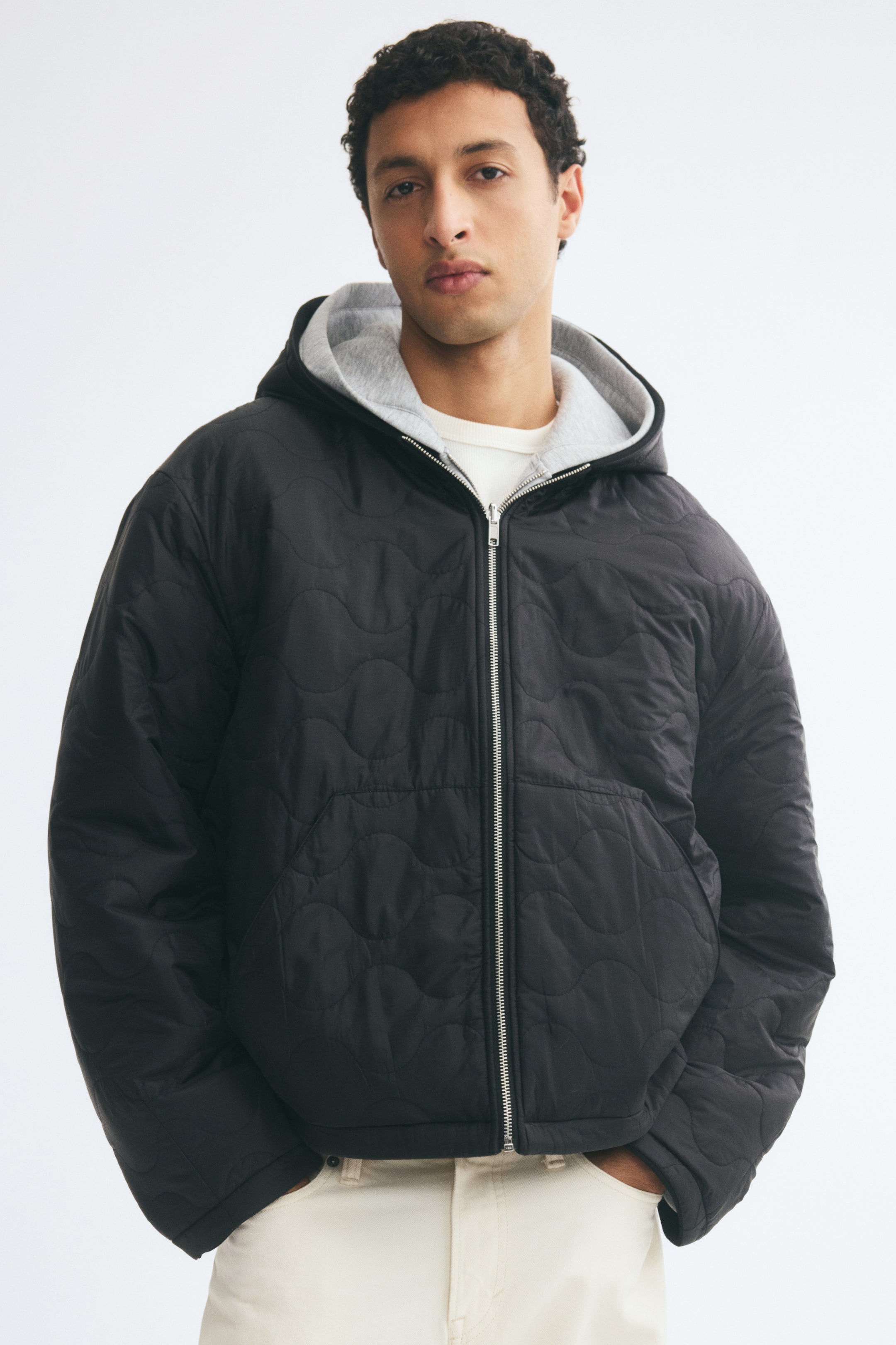Agrandir l'image: Une veste à capuche réversible noire, ornée d’un motif matelassé ondulé, est portée par le mannequin sur un t-shirt blanc et un pantalon clair. La veste présente une fermeture éclair argentée sur le devant, des épaules tombantes et une doublure de capuche gris clair, tandis que le mannequin garde les mains dans les poches avant.