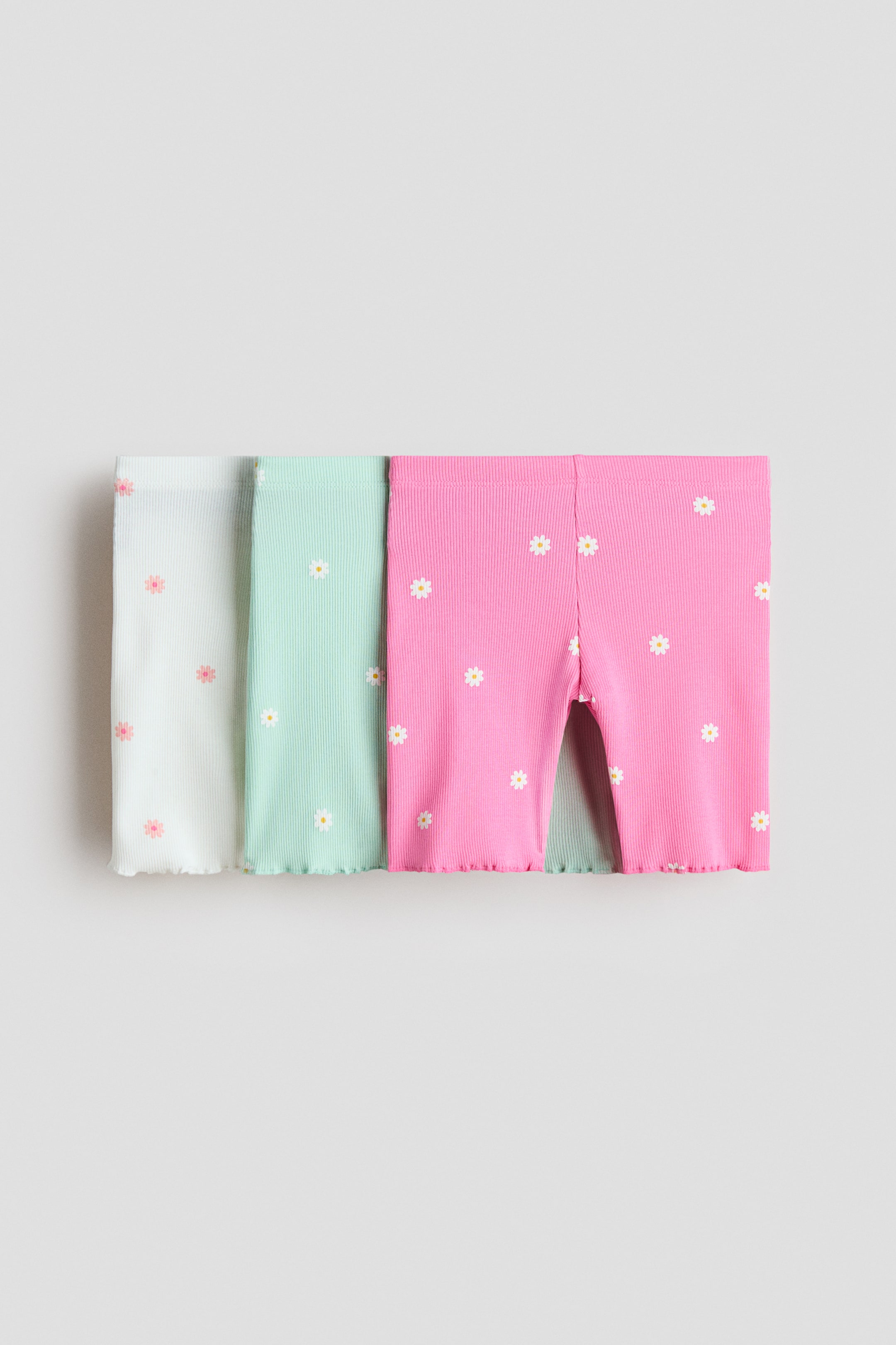 View larger image: Three pairs of ribbed shorts are displayed in a slightly overlapping manner, each showcasing a ruffled leg hem and a daisy pattern. The collection includes a white pair adorned with pink daisies, a mint green pair with white daisies, and a pink pair with white daisies; the pink pair is positioned at the front, highlighting its short length and crotch seam.