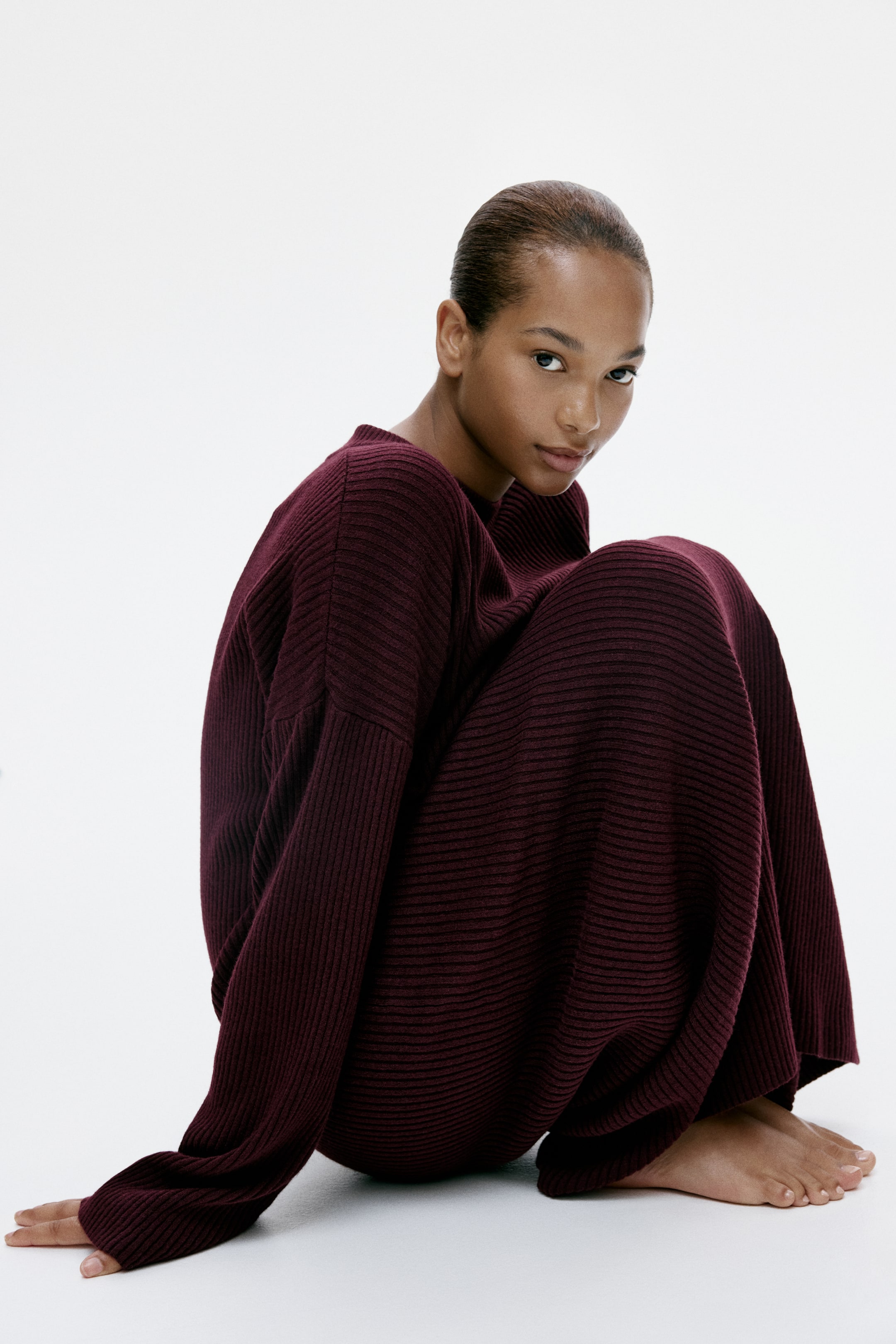 View larger image: A maroon ribbed-knit dress features a round neckline and long, voluminous sleeves with ribbed cuffs, draping loosely over a seated figure to showcase its relaxed fit.