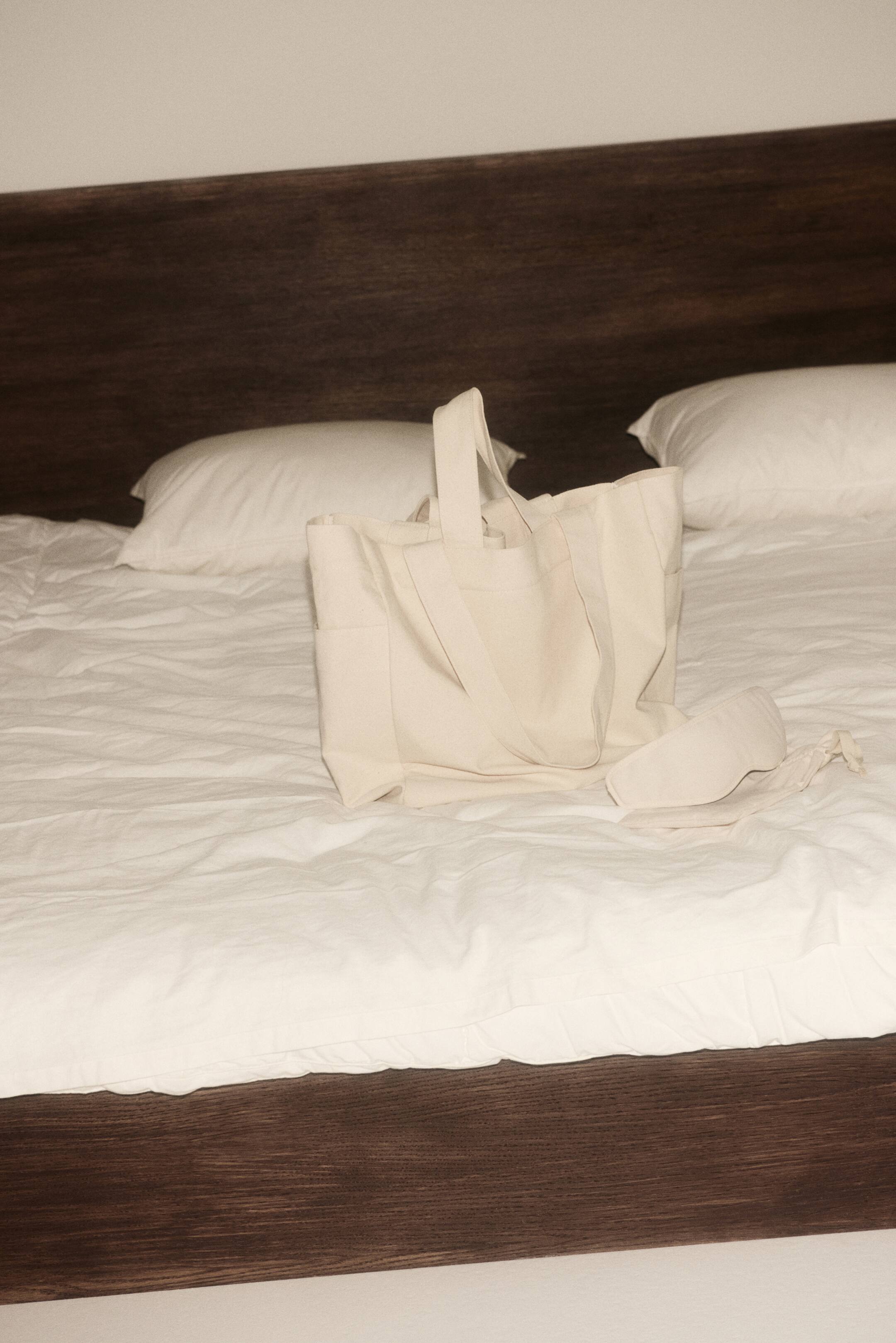 이미지 확대 보기: 크림색 캔버스 토트백이 똑바로 서 있으며, 두 개의 상단 손잡이와 외부 측면 포켓이 특징입니다. 동일한 소재의 수면 안대가 가방 옆에 놓여 있습니다.