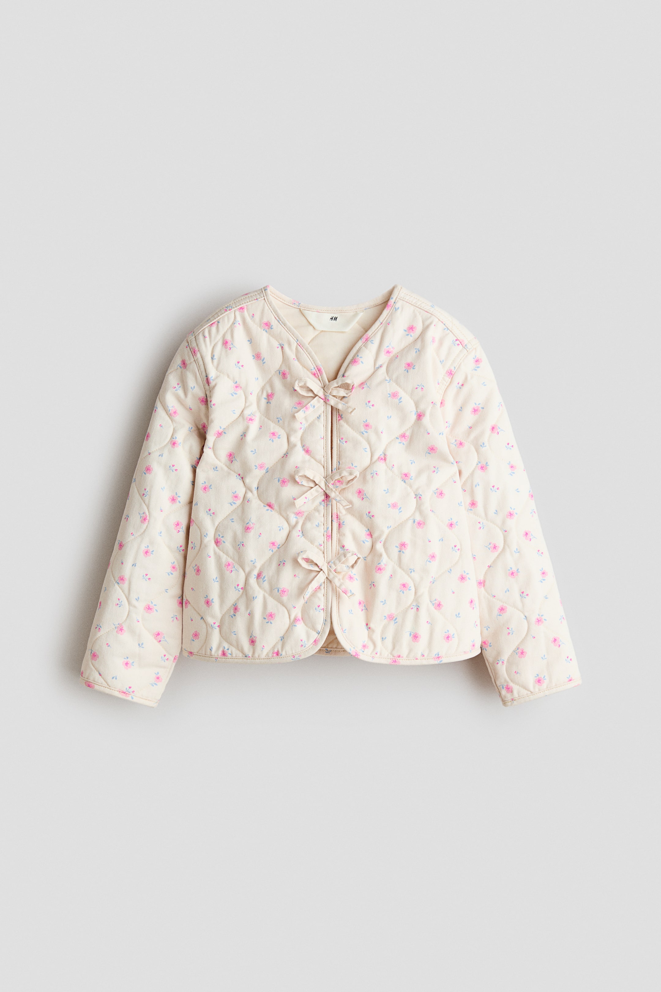 拡大画像を表示: ゆったりとしたフィットのジャケットは、オフホワイトの生地に小さなピンクの花柄の全体的なキルティングパターンが特徴で、前から見られています。Vネックラインでデザインされており、4つの装飾的なリボンが前部を留め、丸みを帯びた裾へとつながっています。